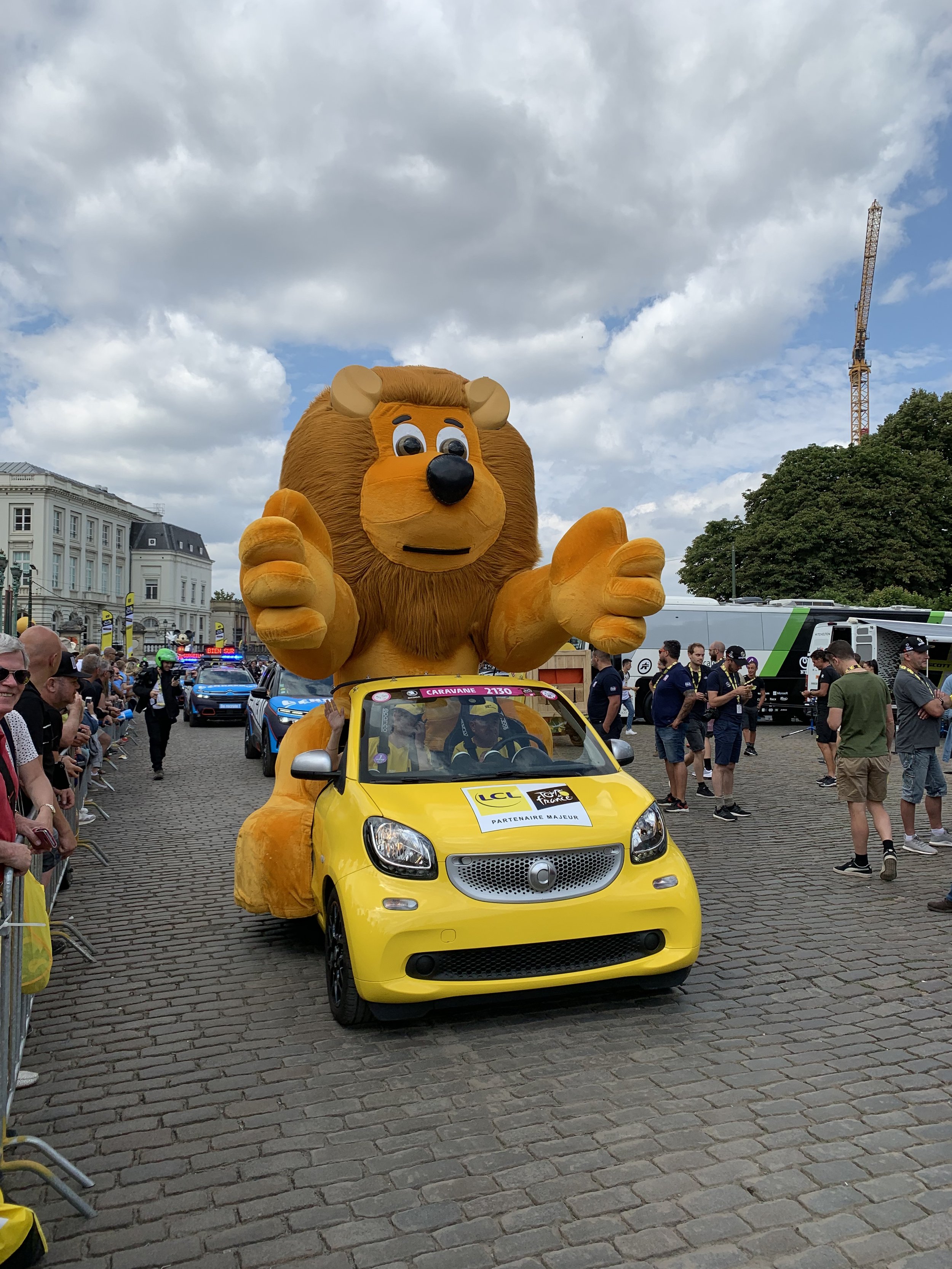 Een grote gele leeuwenkostuum op een openluchtparade, met een klein gele auto eronder. Mensen staan langs de zijkanten van de straat en kijken naar de parade.