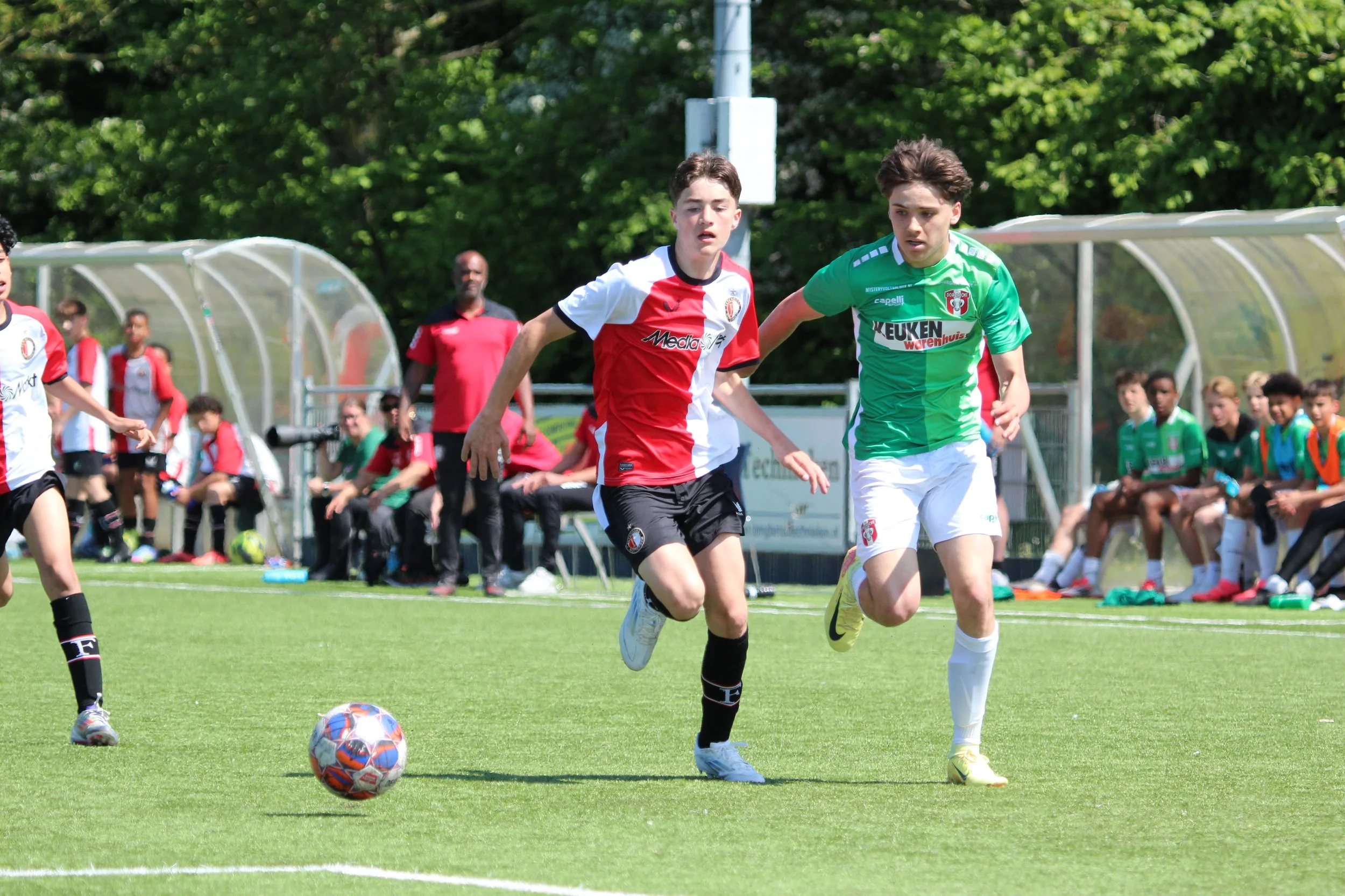 Twee jonge jongens spelen voetbal op een grasveld, een met een rood-wit shirt en de ander met een groen shirt, op een zonnige dag.