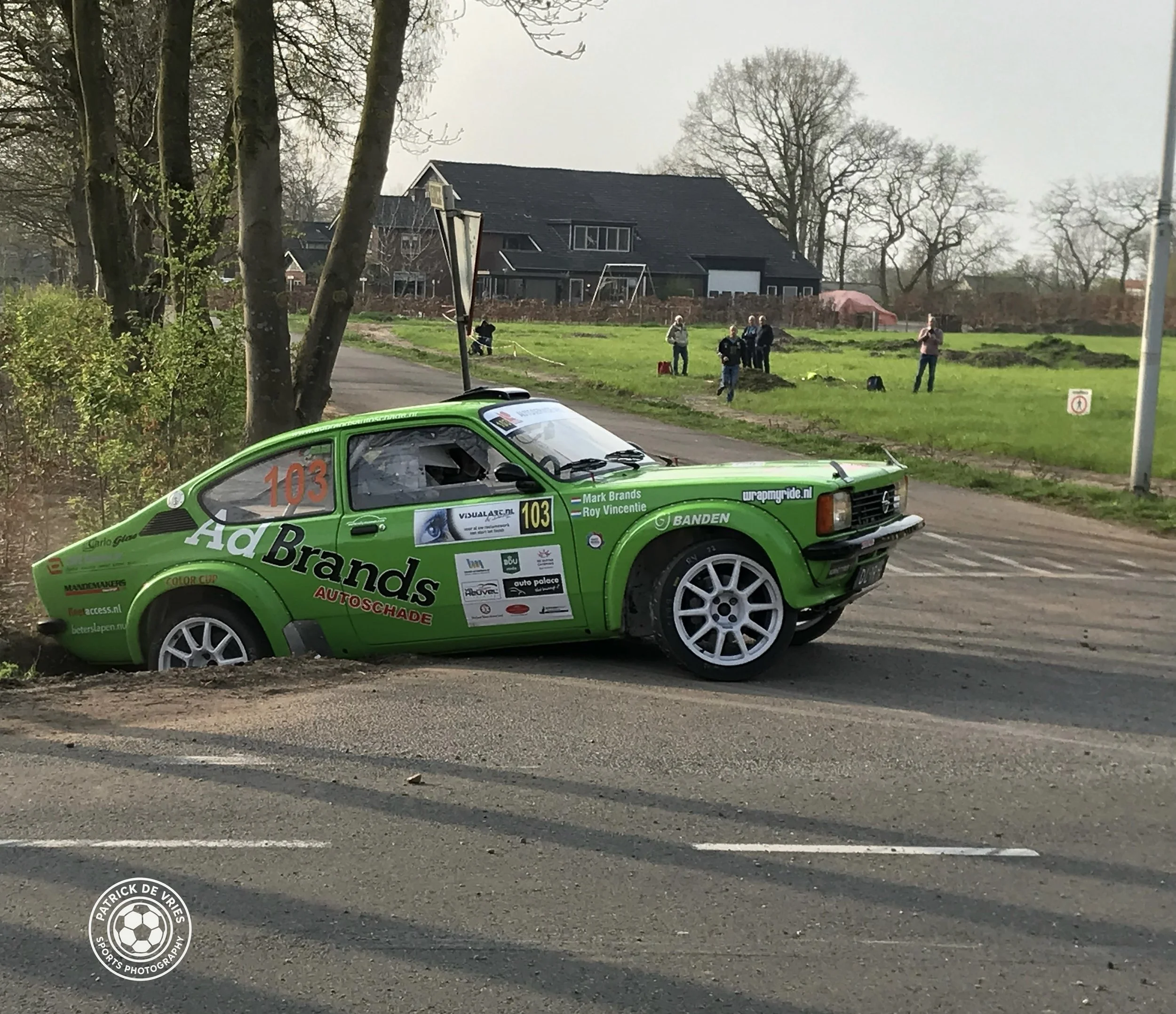Groene raceauto op een weg met een huis en een groep mensen op de achtergrond.