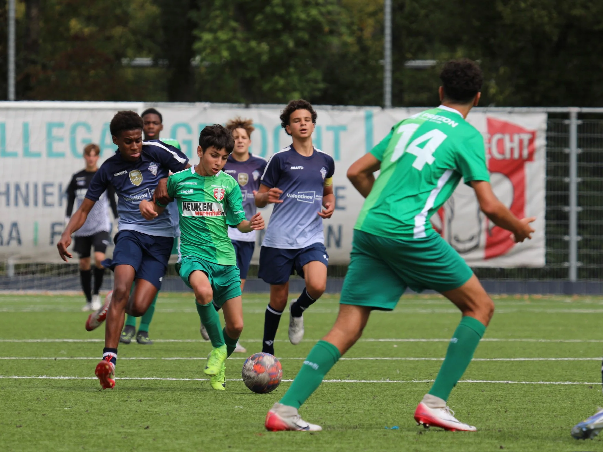 Kleine jongens spelen een voetbalwedstrijd op een grasveld, met enkele spelers in het blauw en andere in groen. Eén jongen in groen probeert de bal te controleren, terwijl de spelers in het blauw proberen hem te verdedigen.