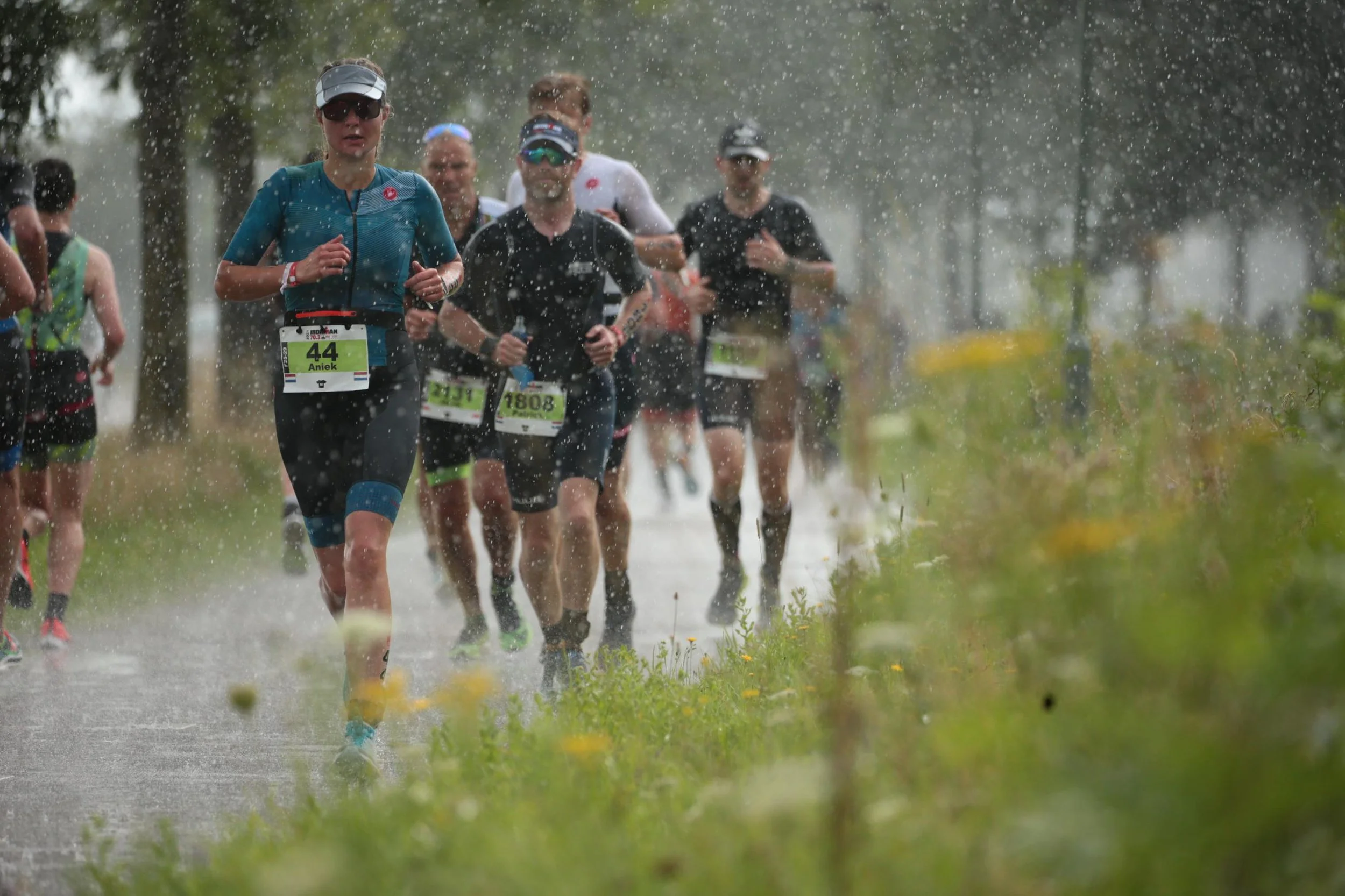 Hardlopende marathonners op een regenachtige route, met regenwalm en potplanten langs de zijlijn.