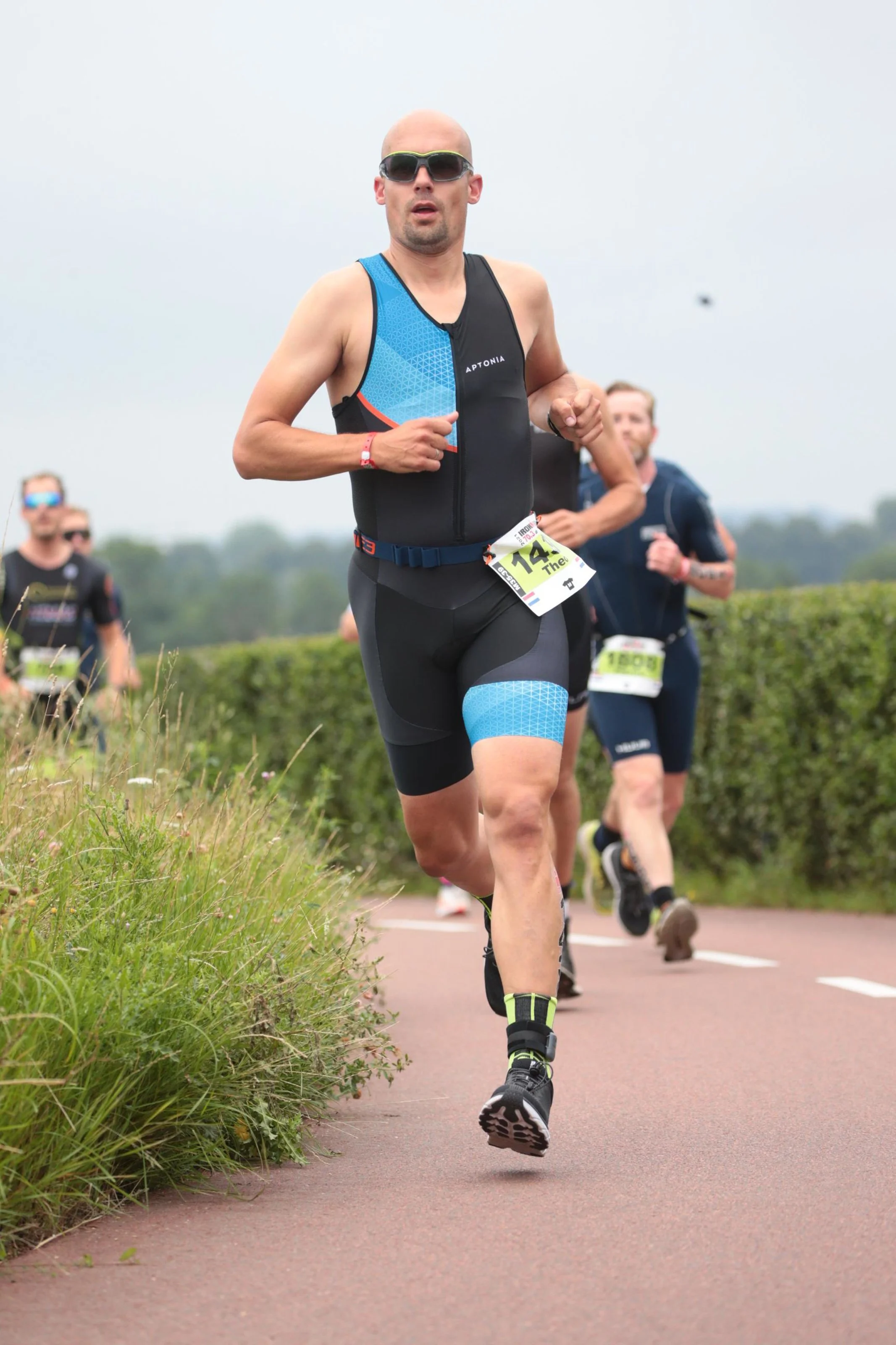 Man die meedoet aan een hardloopwedstrijd, lopend op een weg met andere lopers op de achtergrond, gekleed in sportkleding en sportbril, met een nummer op de voorkant.