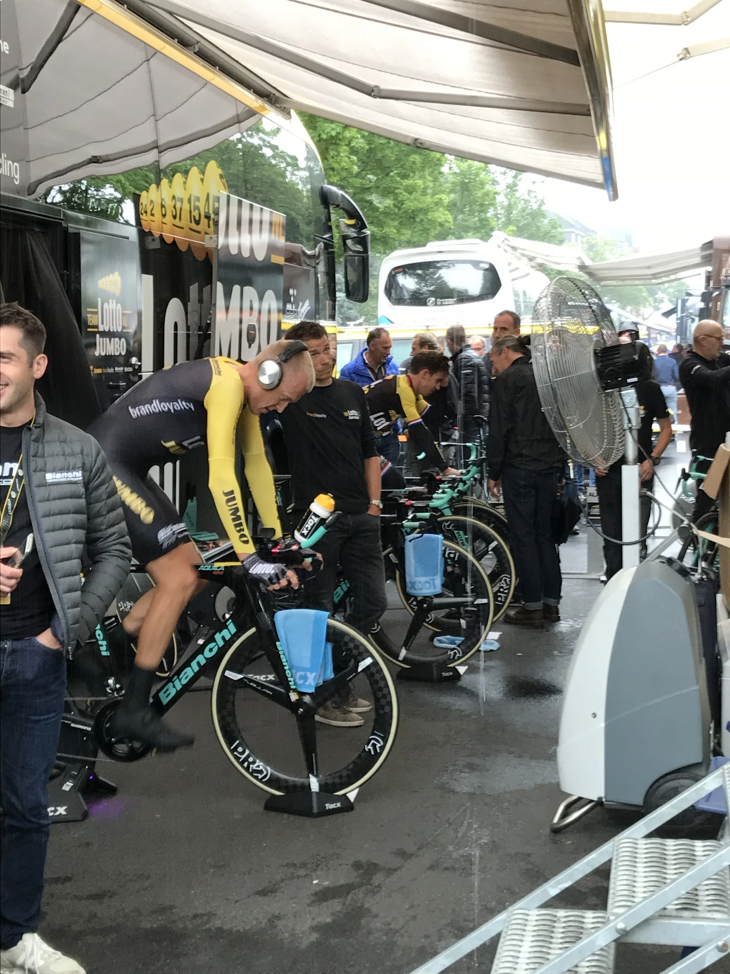 Wielrenners in de startgebied, naast fietsen en digitale monitors, onder een feesttent op een regenachtige dag.