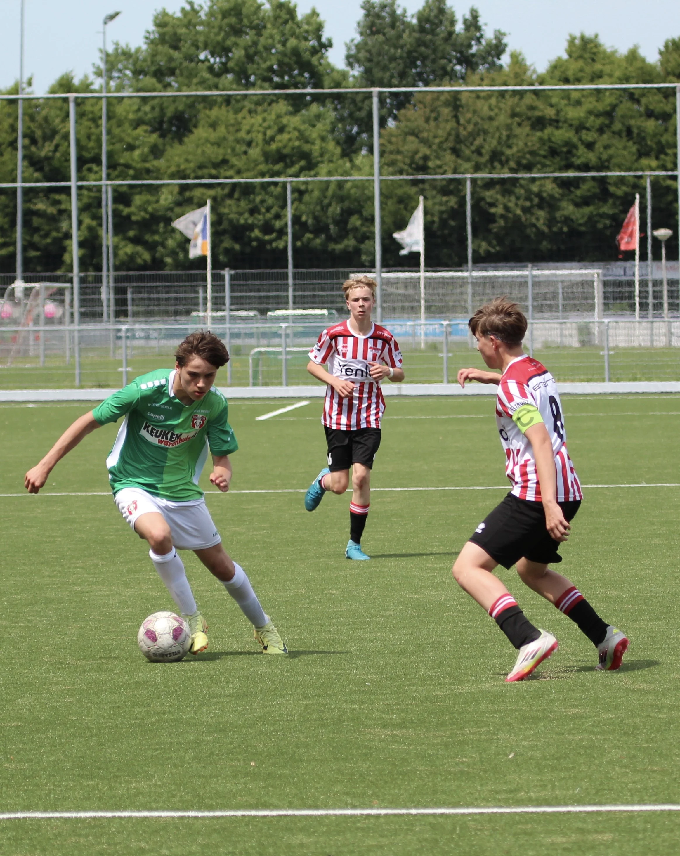 Drie jonge jongens spelen voetbal op een grasveld, met een hek en bomen op de achtergrond.