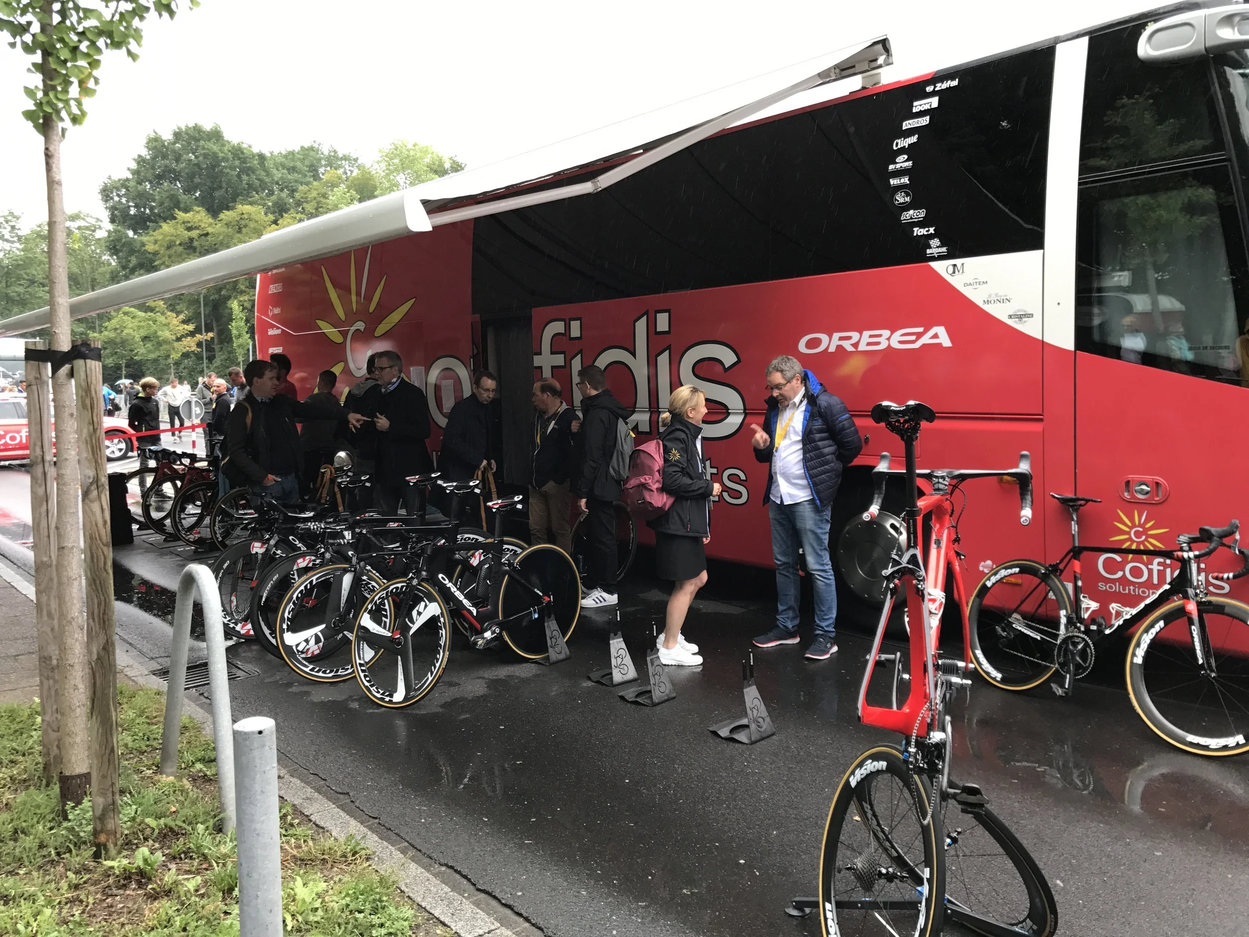 Een groep mensen staat naast een rode fietstruck, met racefietsen uitgestald voor de truck. Het is een natte dag met regen op de grond, en er zijn bomen op de achtergrond.
