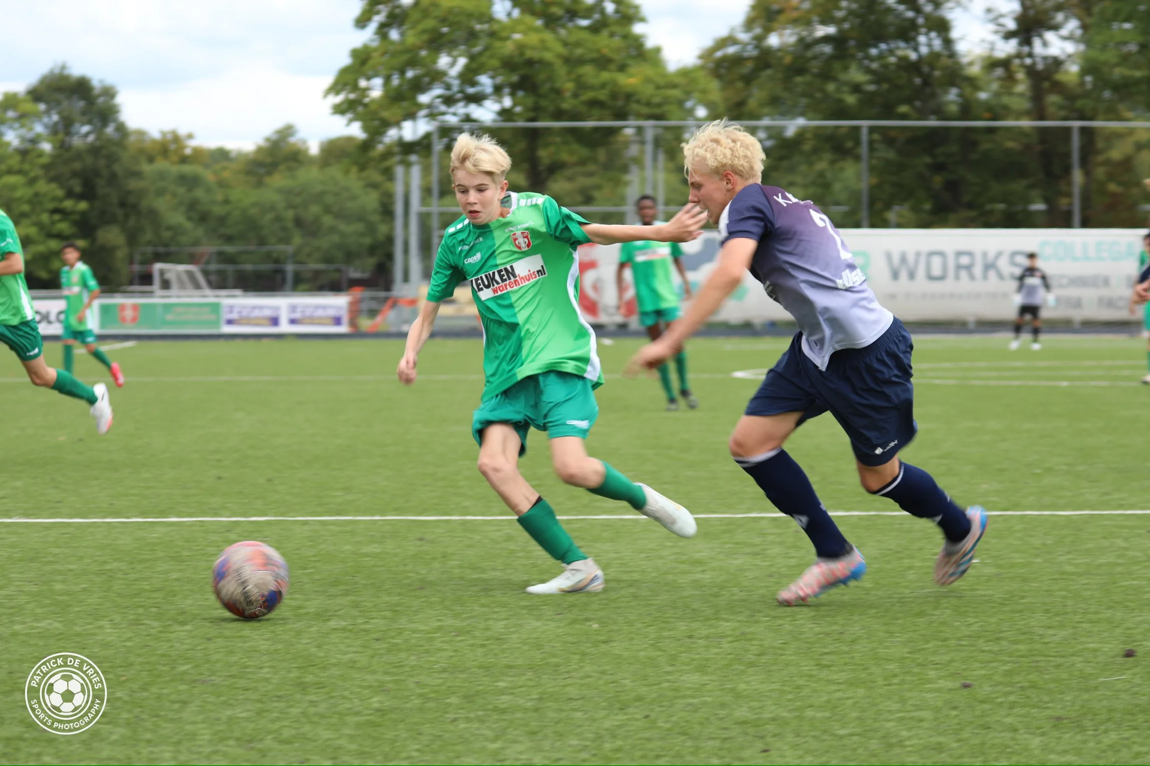 Twee jeugdvoetballers strijden om de bal tijdens een wedstrijd op een grasveld, met andere spelers op de achtergrond en bomen aan de horizon.