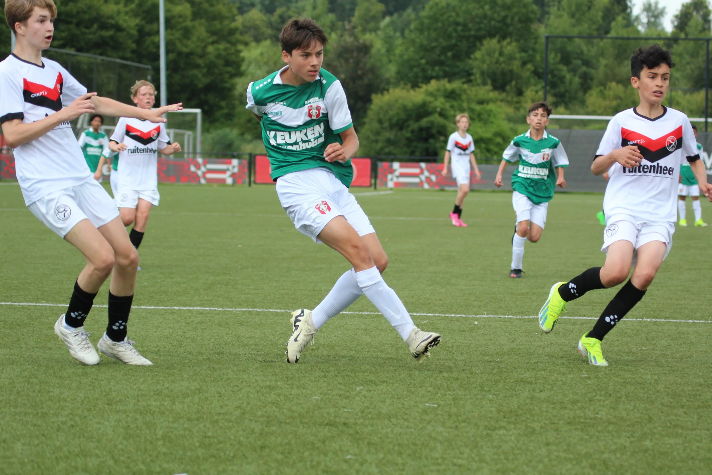 Jongens spelen jeugdvoetbal op een grasveld, enkele jongens in witte truien en enkelen in groene truien, onder een bosrijke achtergrond.