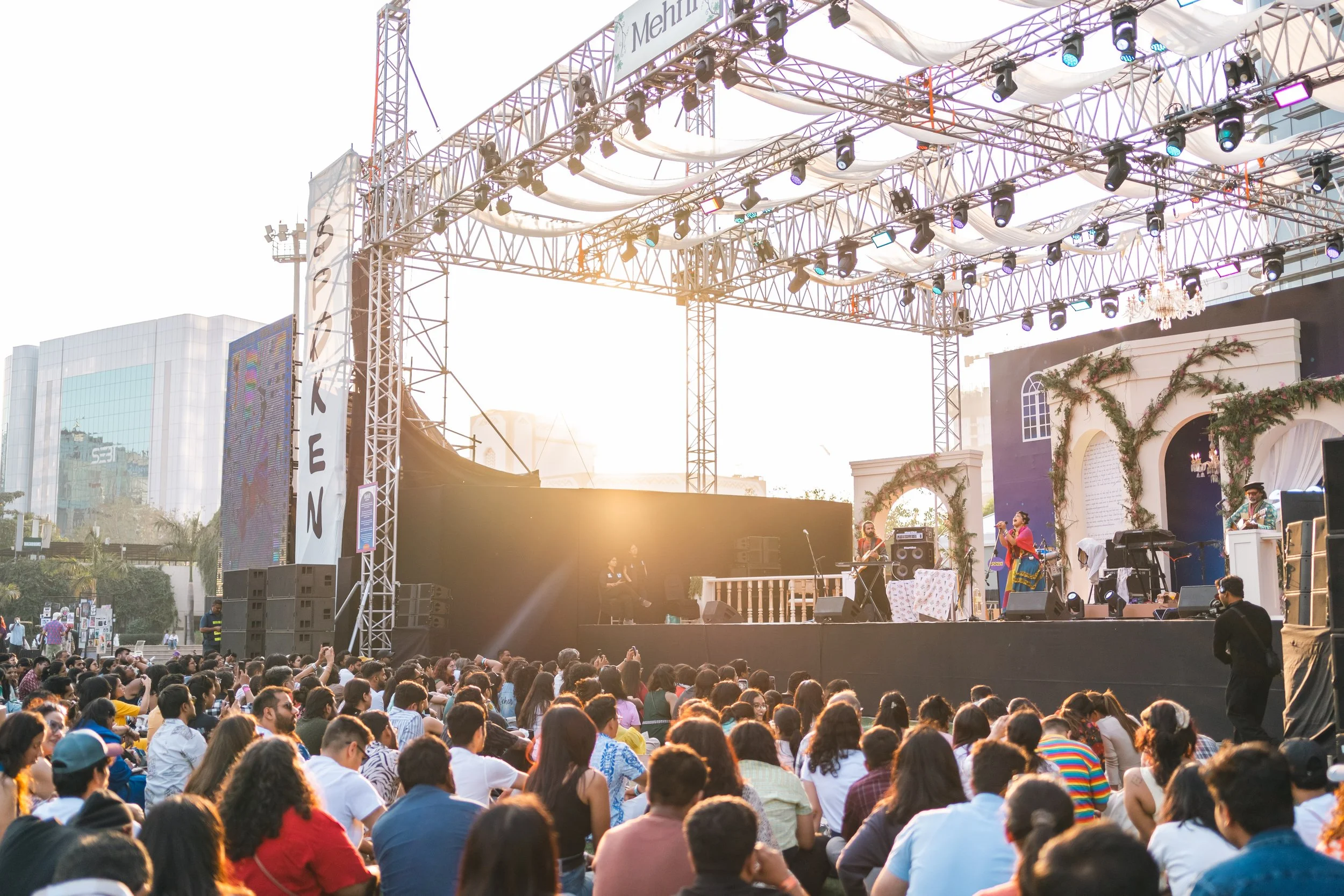 A large crowd is gathered in front of a stage, with a band performing under a metal and white draped canopy. The stage is decorated with greenery and a chandelier, with sunlight illuminating the scene from the background. Tall buildings and a digital screen are visible in the distance.