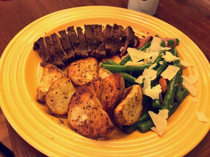 Steak, roasted potatoes, green beans with cheese, and garlic on a yellow plate.