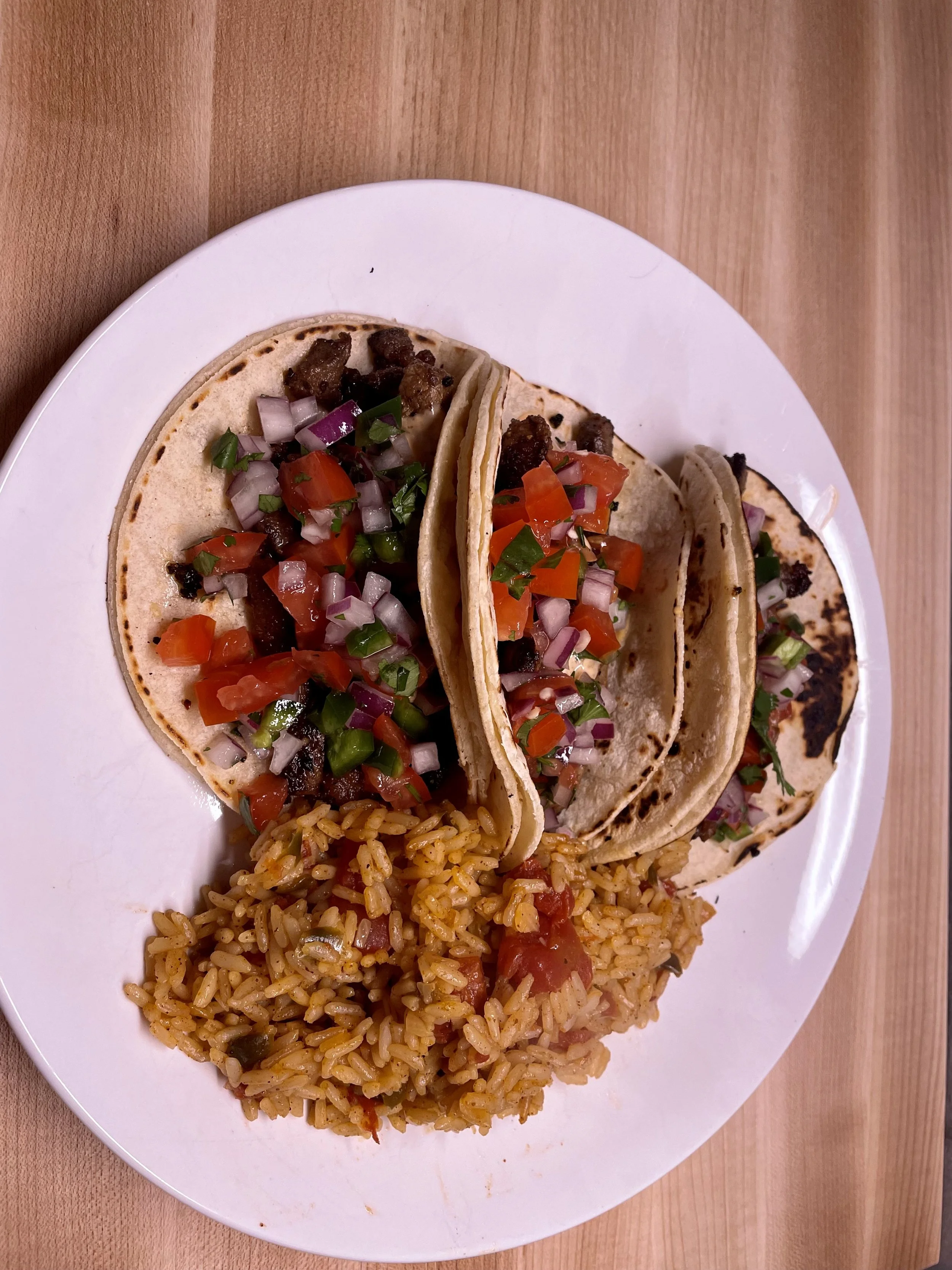 Three street tacos with diced tomatoes, onions, cilantro, and meat, served with a side of Mexican rice on a white plate.