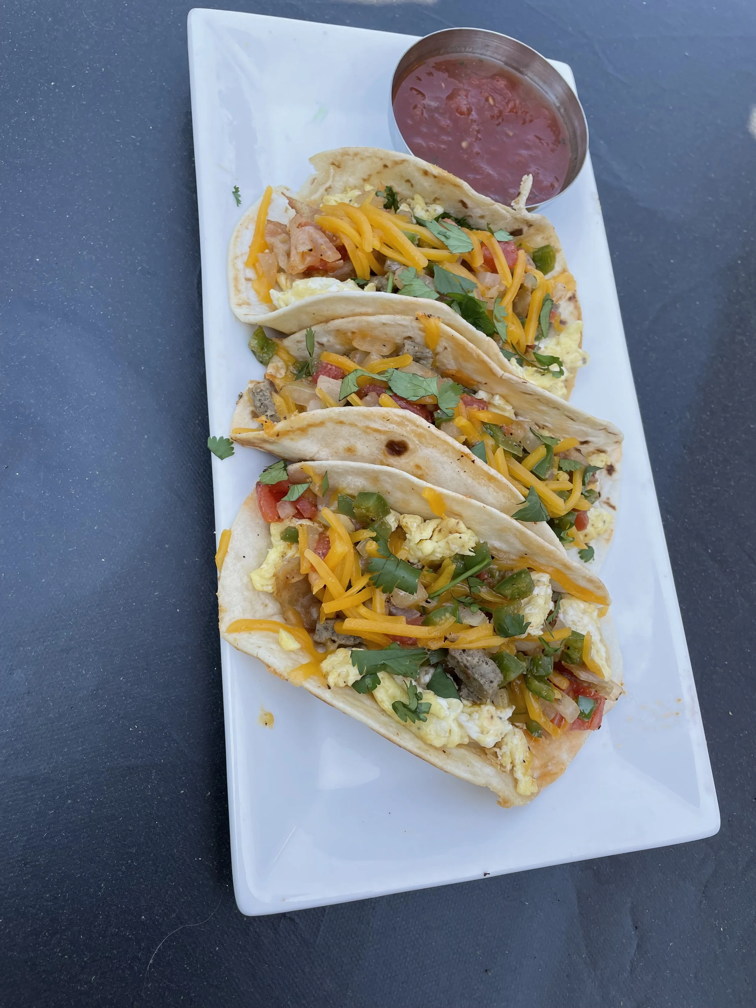 Three tacos filled with scrambled eggs, shredded cheese, beef, tomatoes, onions, green peppers, and cilantro, served on a white rectangular plate with a small cup of red salsa.