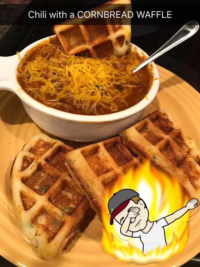 A bowl of chili topped with shredded cheese and a cornbread waffle, with additional cornbread waffles on a wooden plate, and a cartoon character dabbing with flames around him.