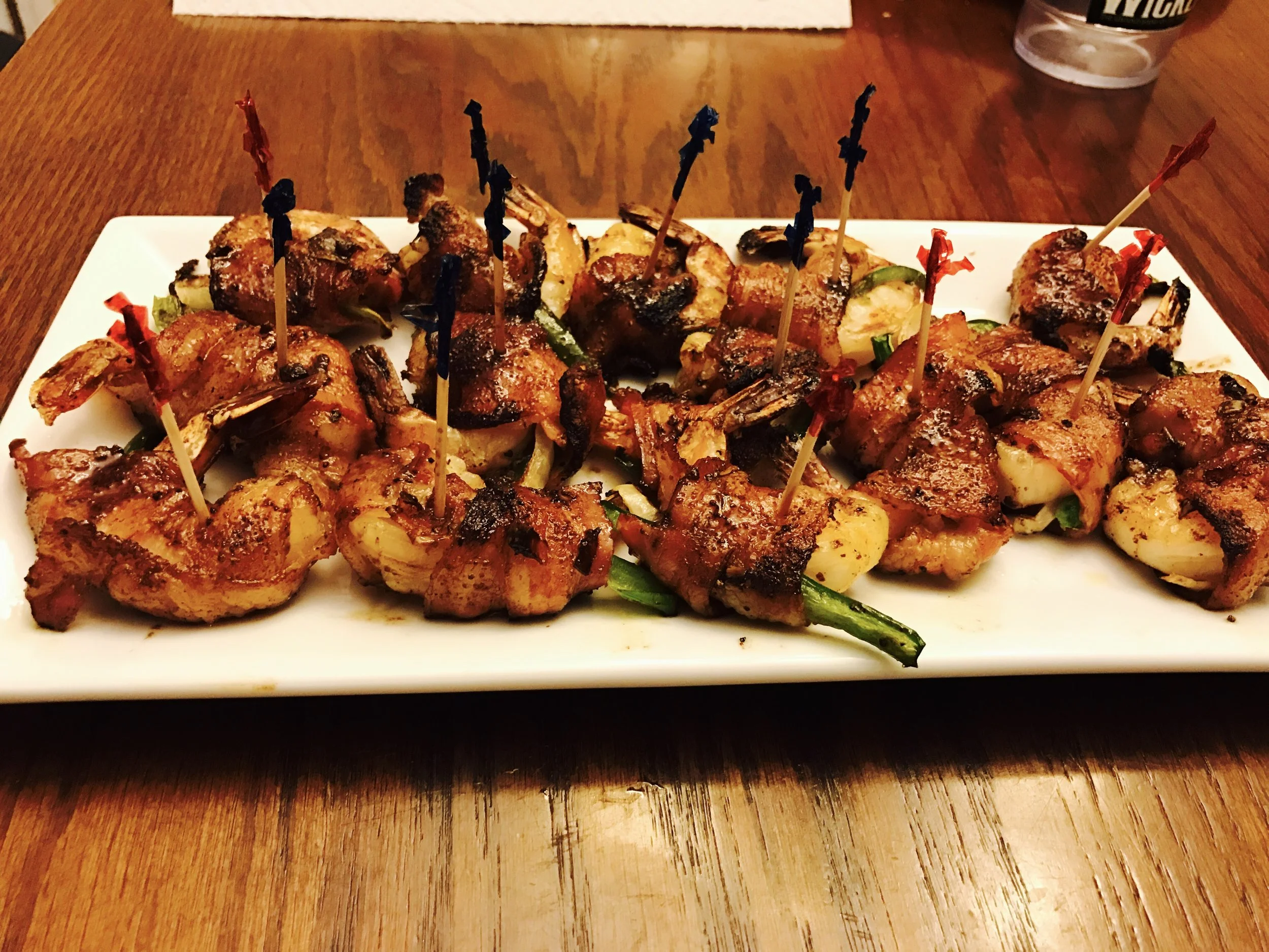A rectangular white platter filled with grilled meats and vegetables, topped with decorative toothpicks, on a wooden table. A drink cup is visible in the background.
