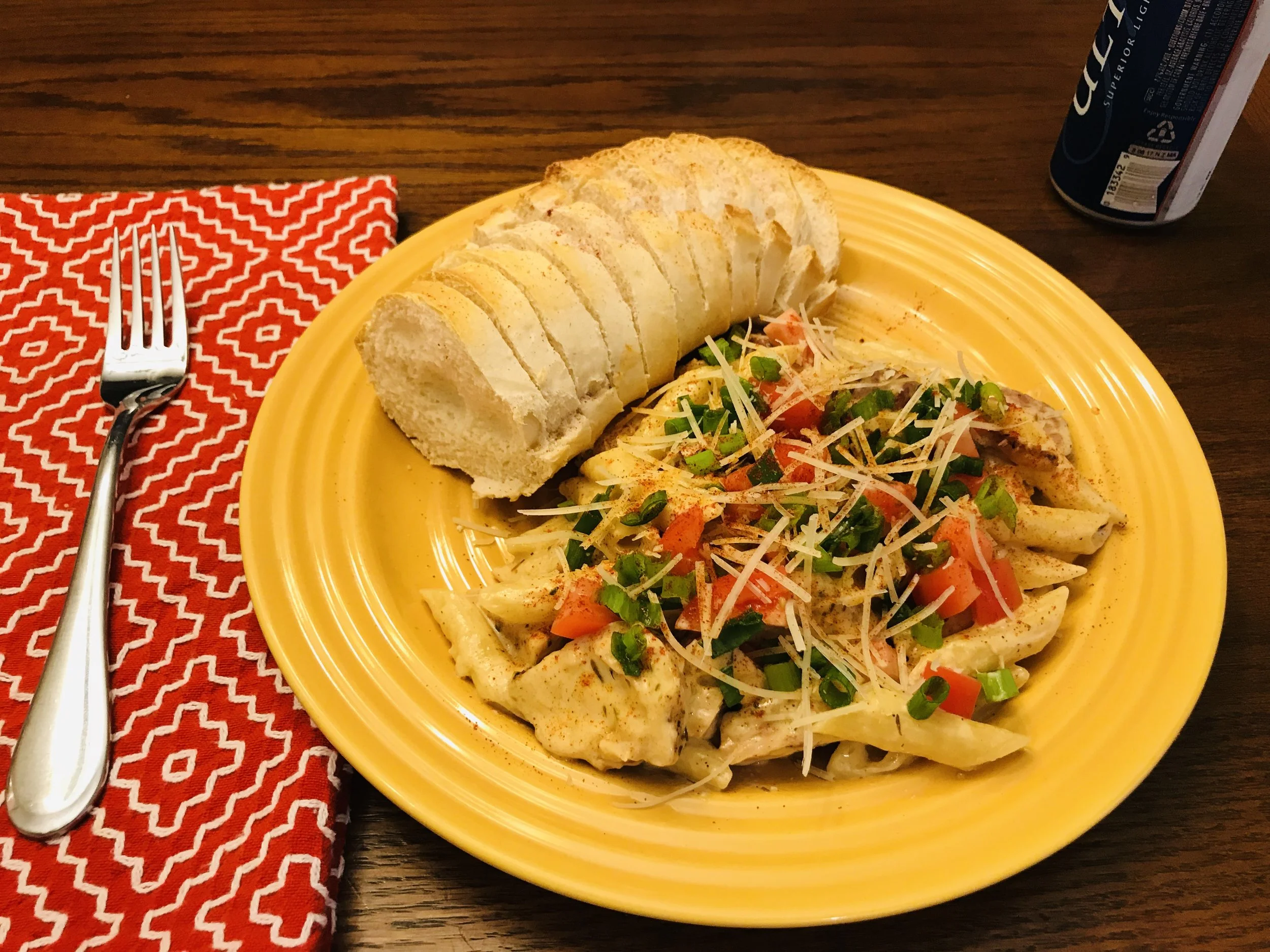 A plate of creamy pasta topped with chopped green onions, shredded cheese, and red peppers, served with sliced bread on a yellow plate. A fork rests on a red patterned napkin beside the plate, and a soda can is partially visible in the background.
