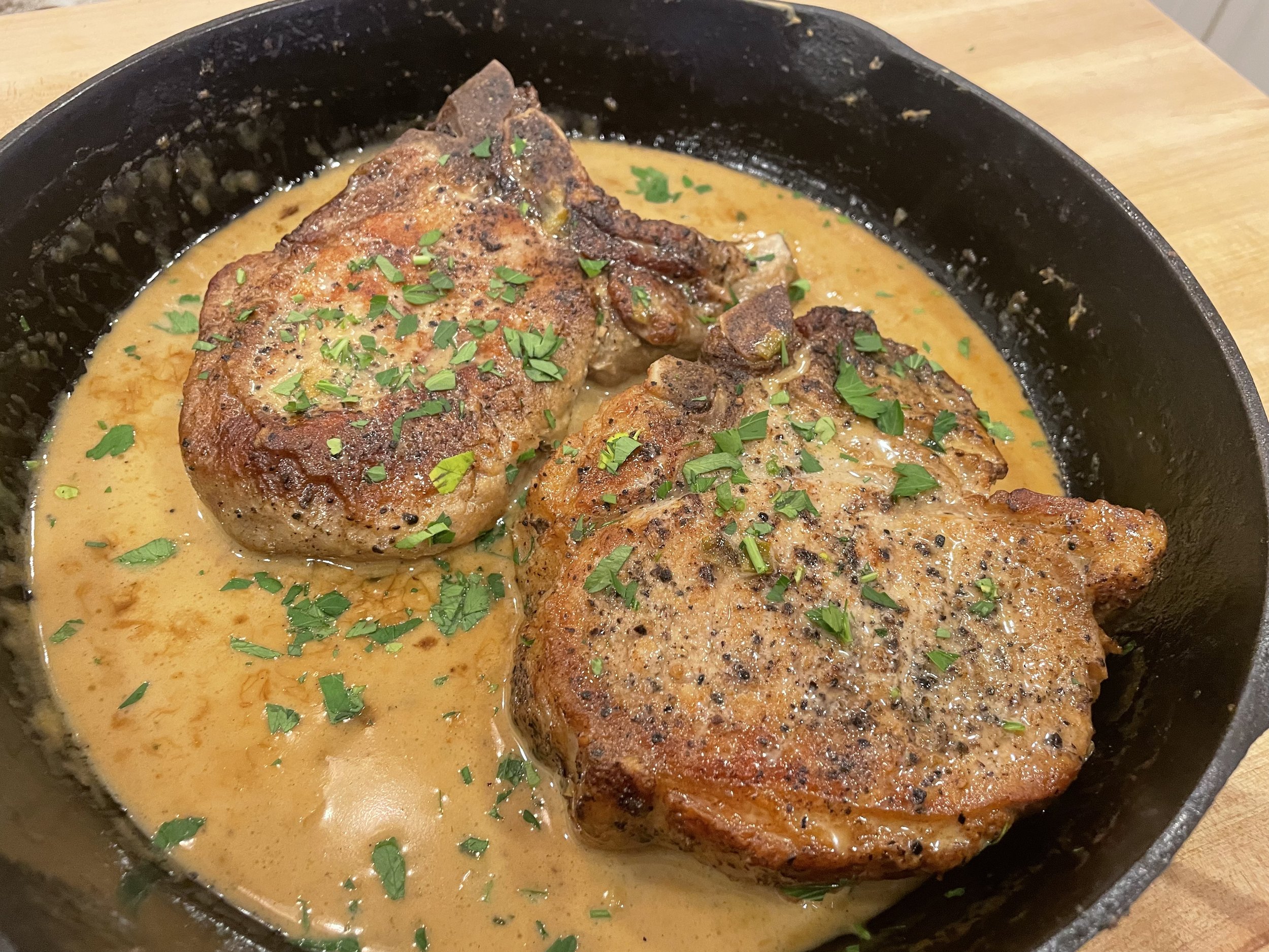 Two seasoned pork chops cooked in a creamy sauce, garnished with chopped parsley, in a black skillet.