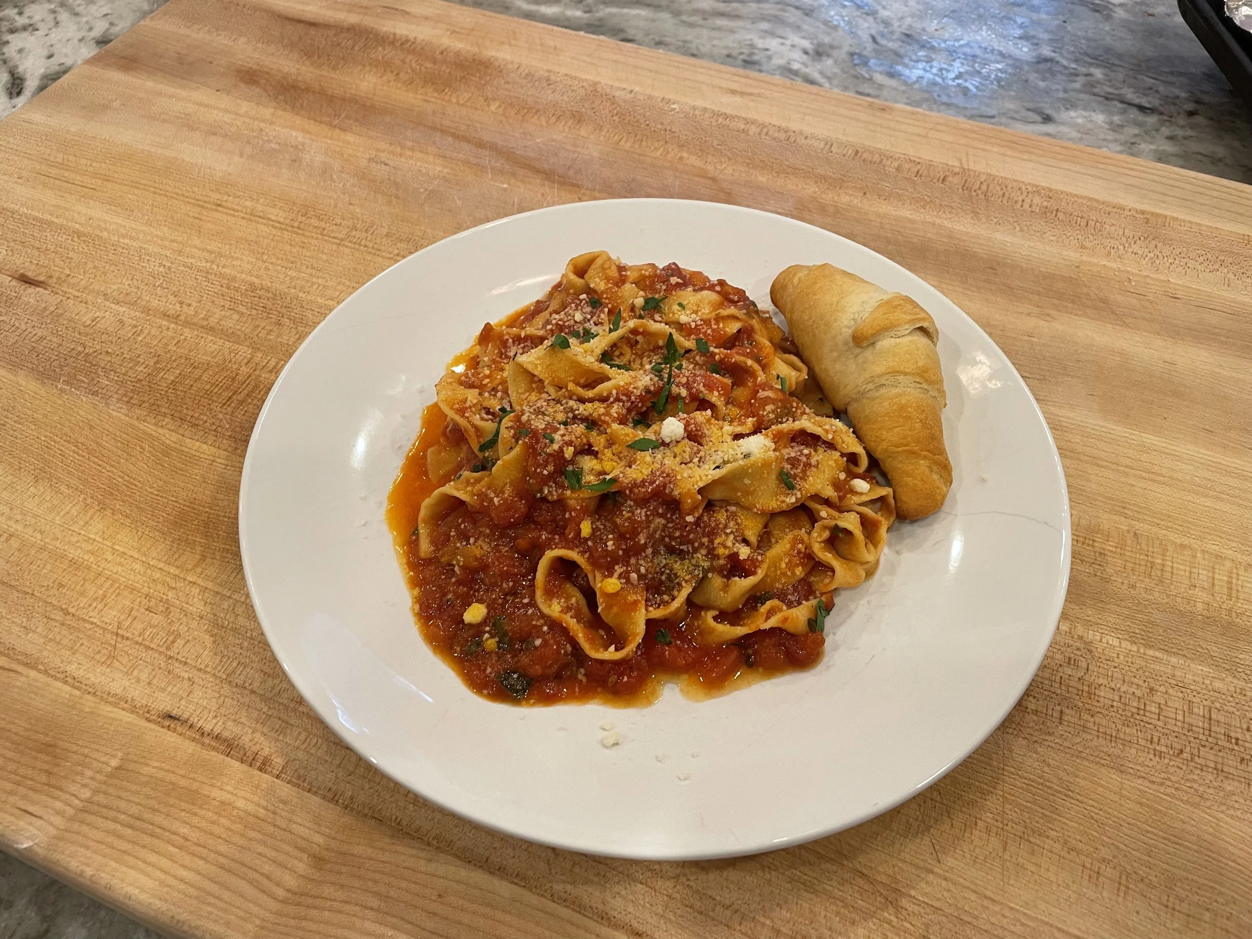 Plate of pasta with tomato sauce, topped with grated cheese and herbs, served with a bread roll on a wooden surface.