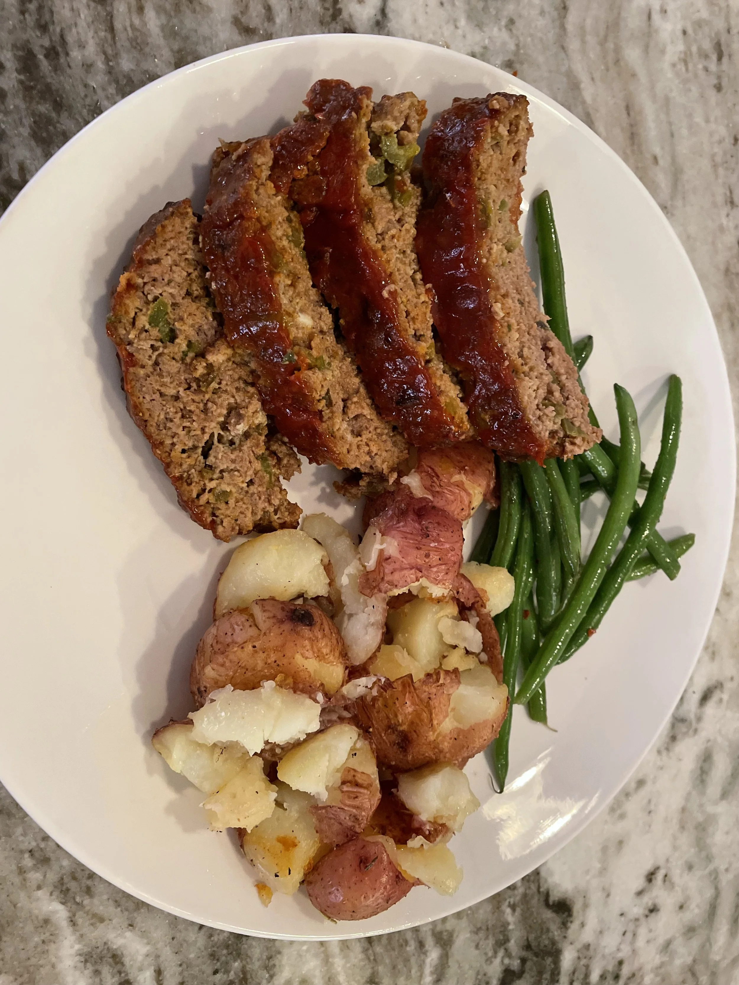 Baked meatloaf slices with ketchup, green beans, and roasted potatoes on a white plate.
