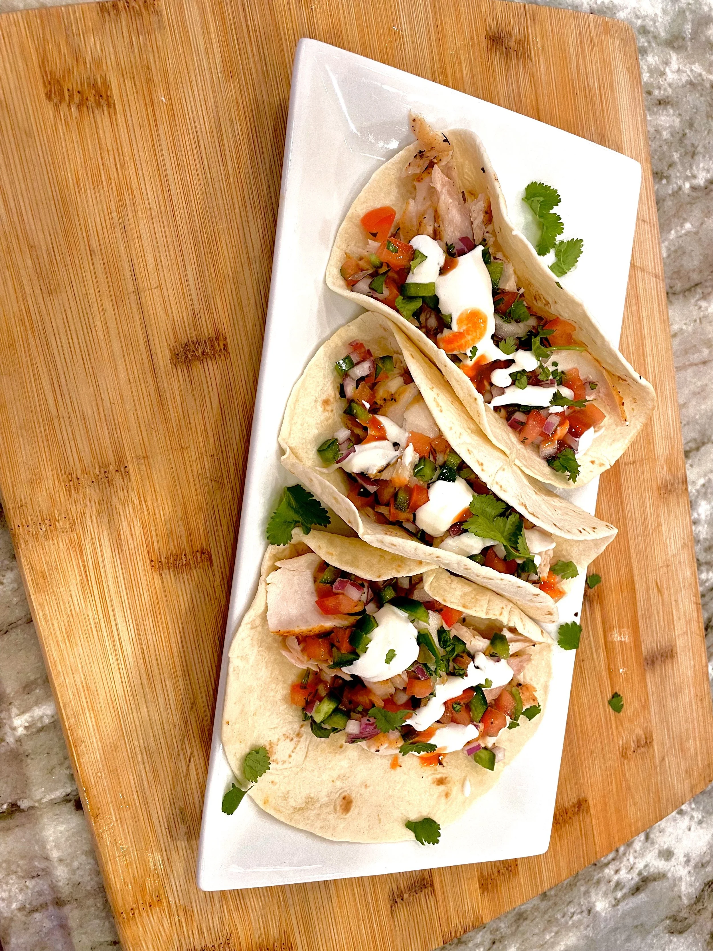 Three tacos topped with chopped vegetables, cilantro, sour cream, and a boiled egg slice on a white rectangular plate on a wooden surface.
