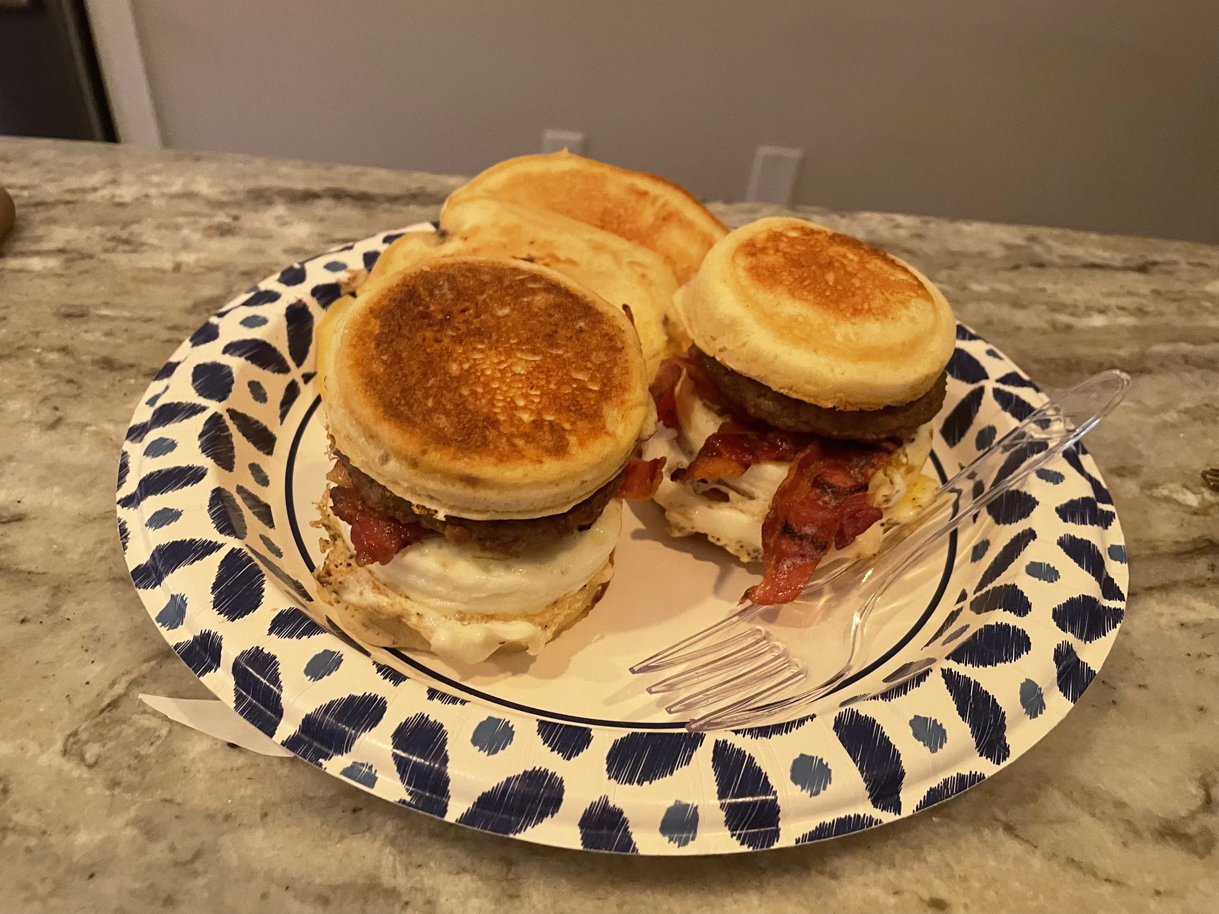 Plate of breakfast sliders with fried eggs, bacon, sausage patty, and toasted English muffins, placed on a decorative paper plate on a marble countertop.