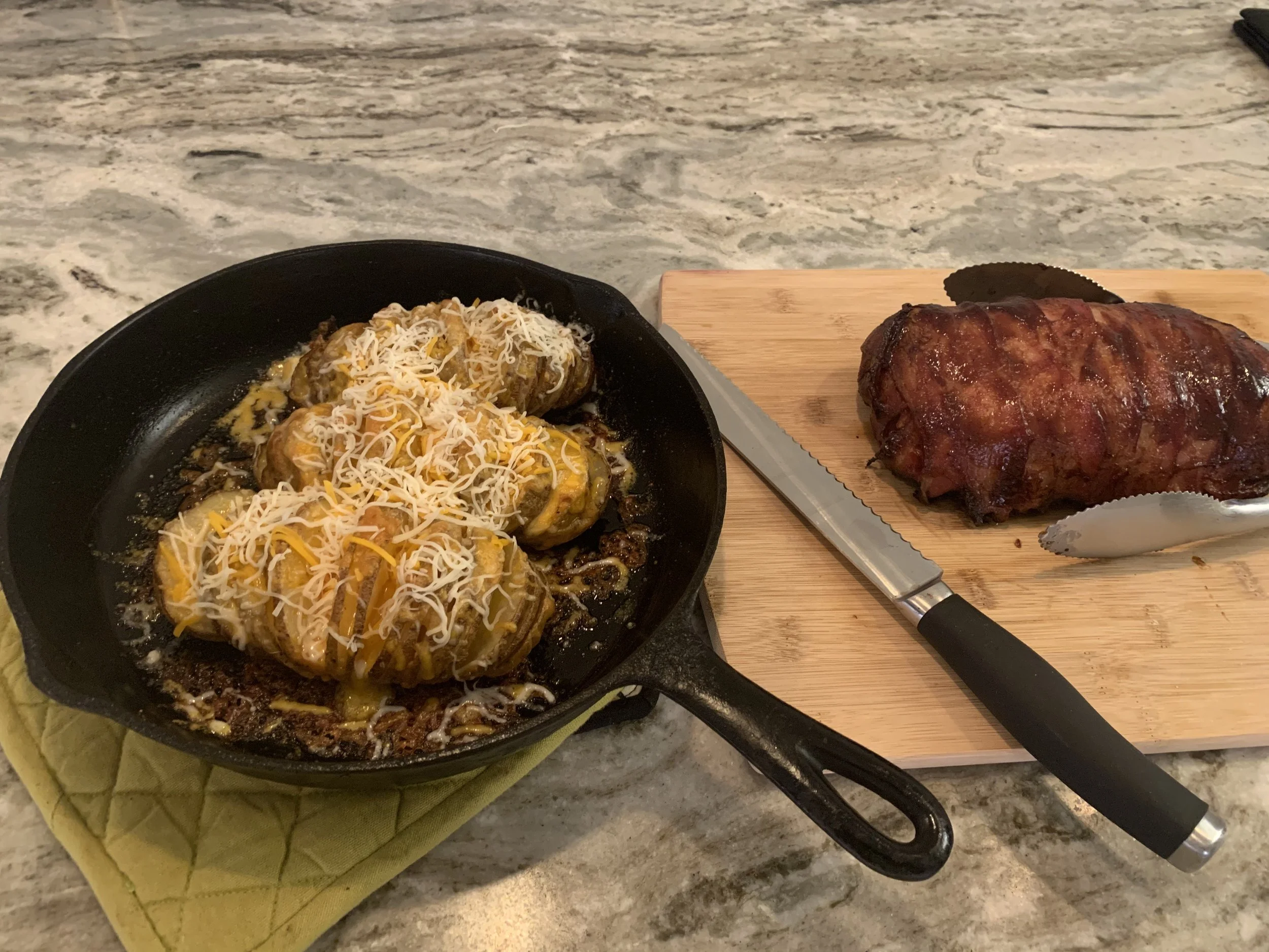 Cast iron skillet with cheesy stuffed jalapeño poppers on a yellow potholder next to a wooden cutting board with a barbecue pork loin and a knife on a marble countertop.