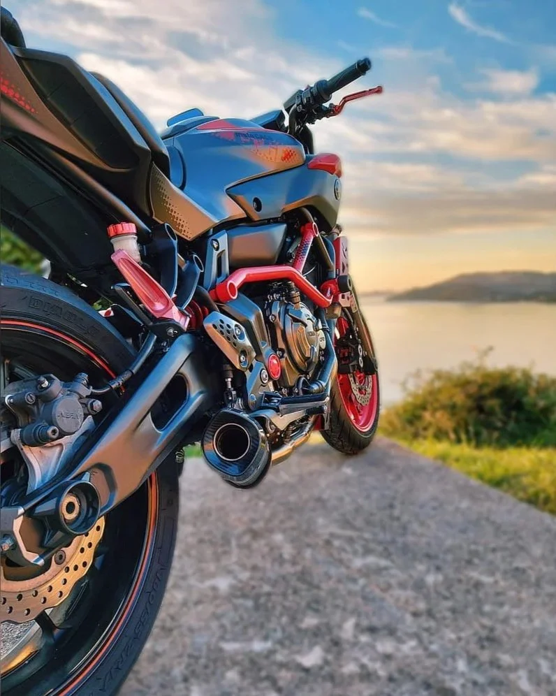 Sportieve motorfiets met zwarte en rode elementen, geparkeerd langs een weg met uitzicht op water en landschap onder een deels bewolkte hemel.