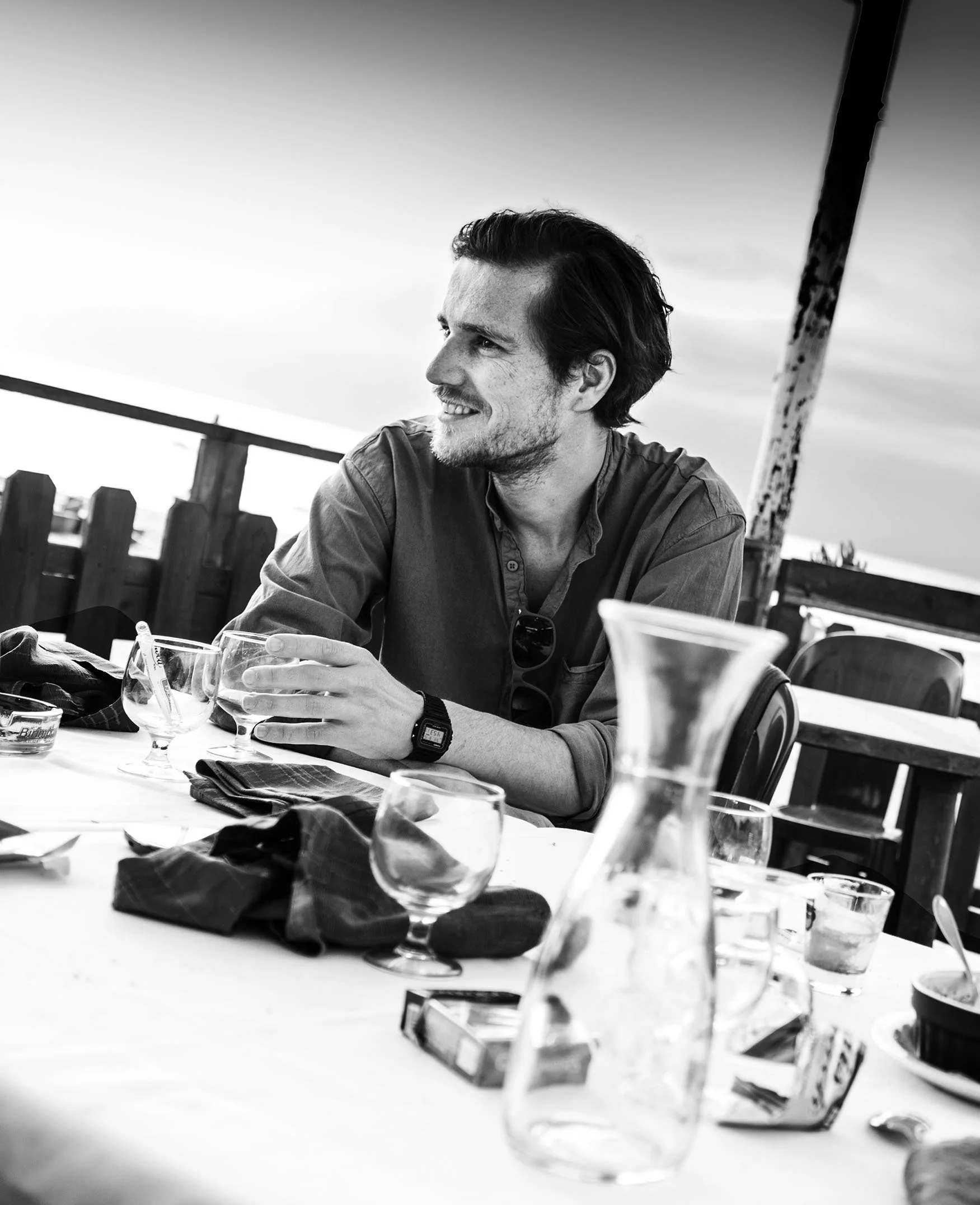 Black and white photo of a smiling man with dark hair and stubble, sitting at a table with various beverage glasses and packages, outdoors with a wooden fence and sky in the background.