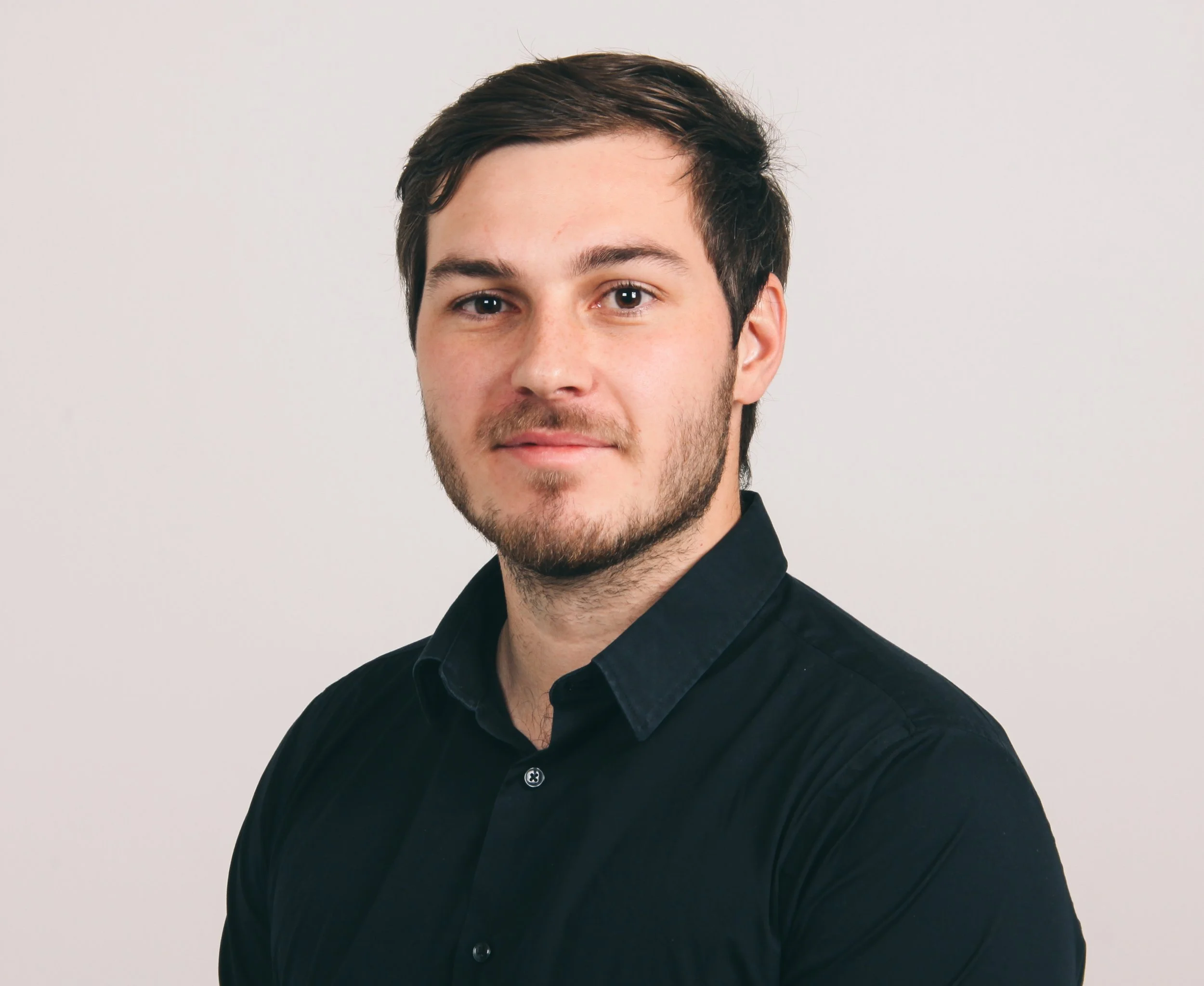 Portrett av en ung mann med mørkt hår, skjegg og i svart skjorte, stående mot en lys bakgrunn.