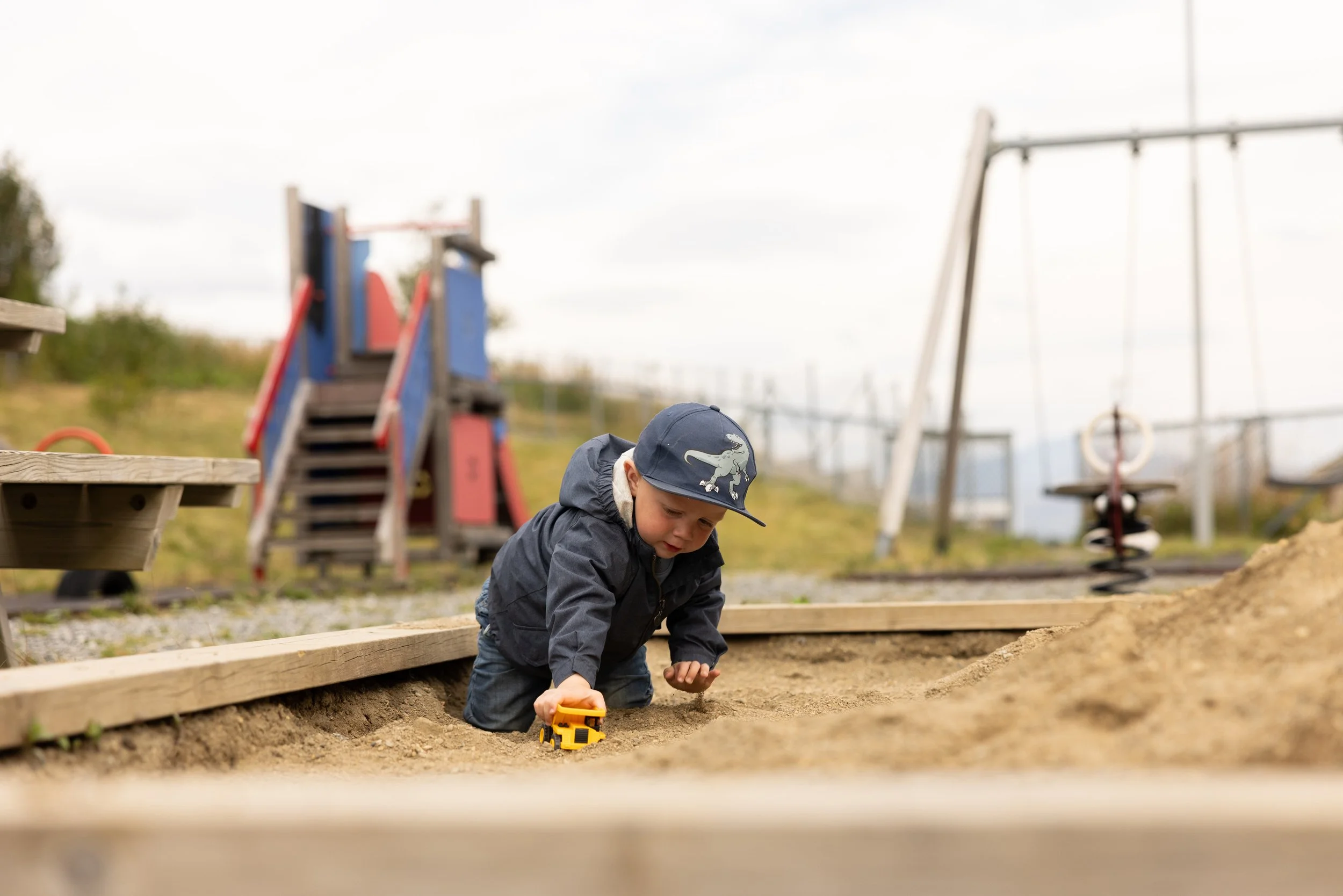 En unge gutt i mørk jakke og caps leker i sandkassen på lekeplassen.