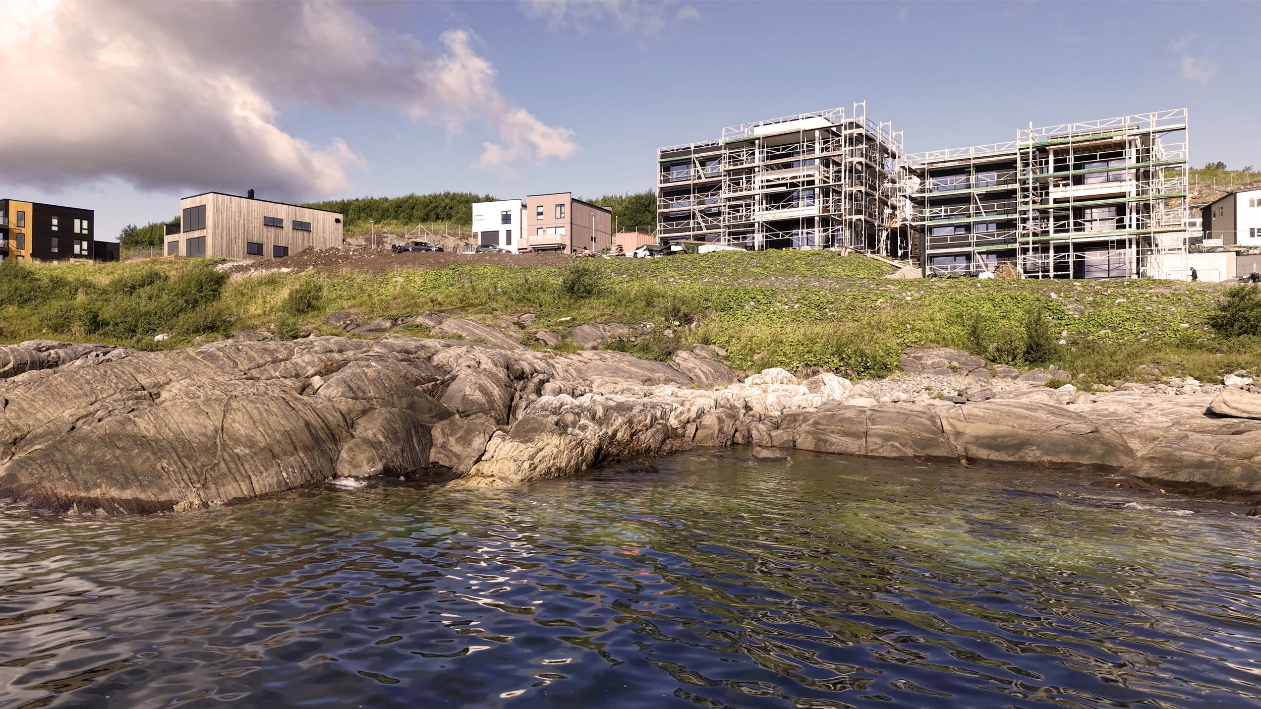 Bygninger under bygging langs en kystlinje med steiner og vann i forgrunnen, solskinn og noen skyer på himmelen.