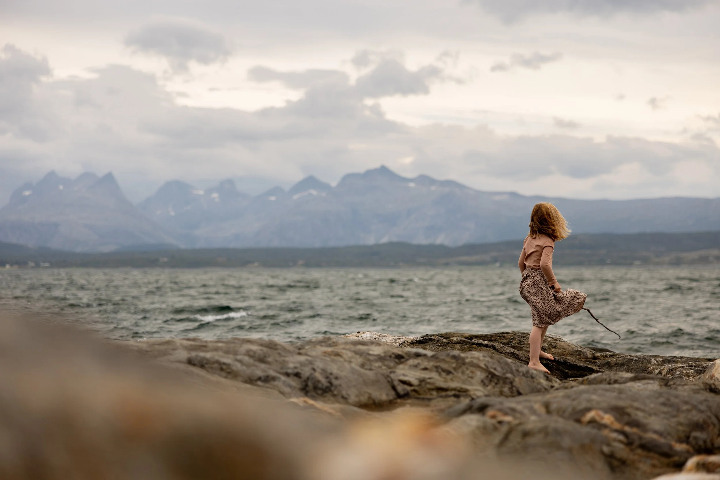 En liten jente står på steiner ved sjøen med fjell i bakgrunnen, under en overskyet himmel.
