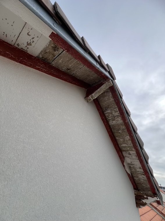 Partie supérieure d'un mur de bâtiment avec des tuiles de toit en arrière-plan, montrant des morceaux de bois ou de soffite endommagés ou usés, sous un ciel nuageux.