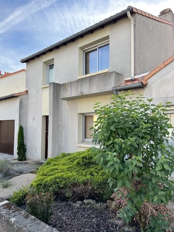 Une maison à deux étages avec un jardin devant, comprenant un arbuste et un buisson vert, et une façade en pierre ou en béton.