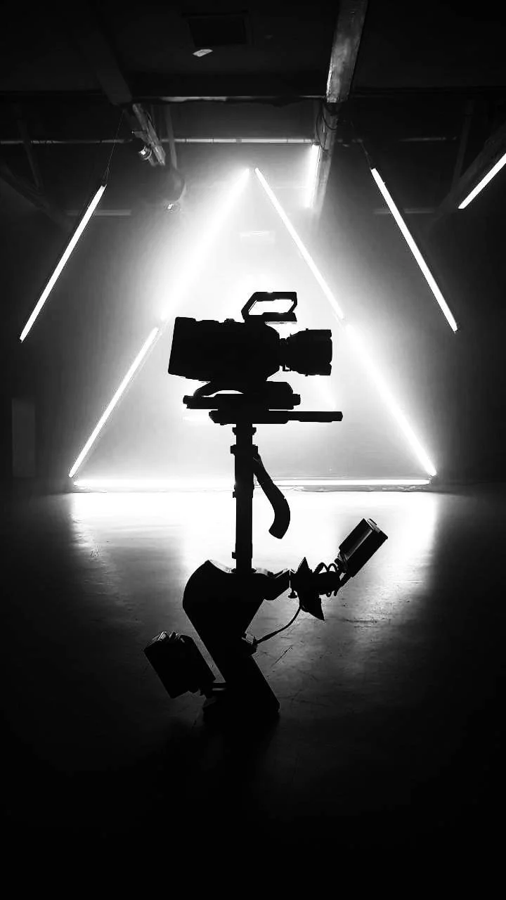 Silhouette einer Videokamera auf einer Steadicam vor leuchtenden diagonalen Lichtstreifen in einem dunklen Raum.