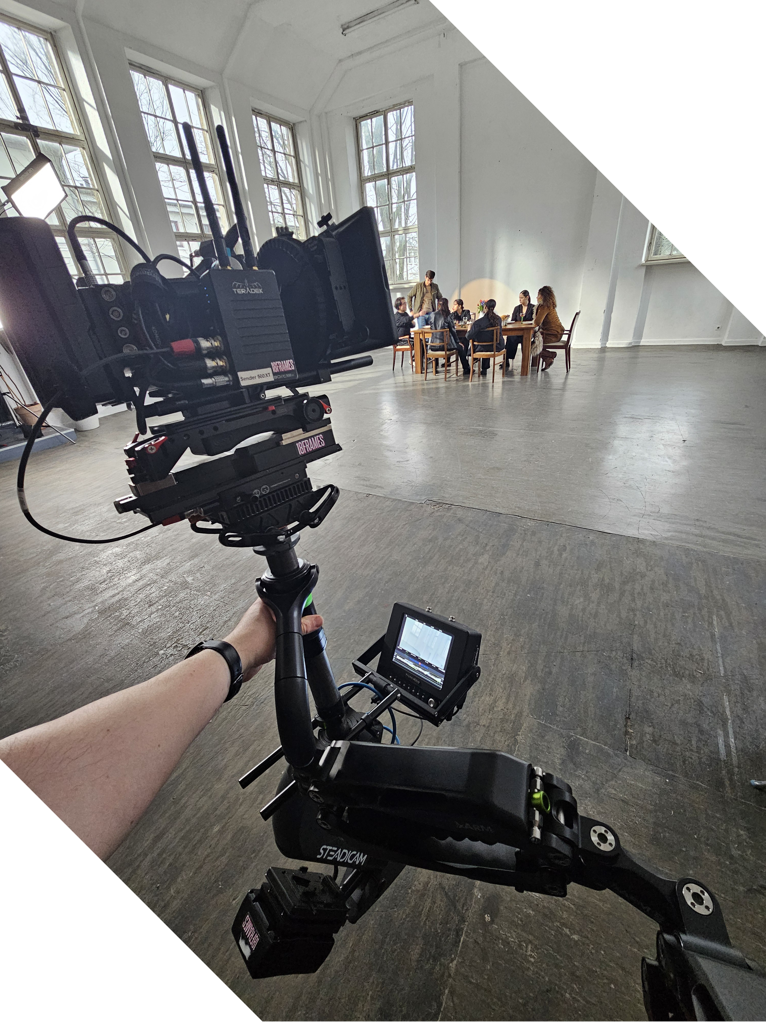 A video camera on a camera rig filming a group of people sitting around a table in a bright, spacious room with large windows.