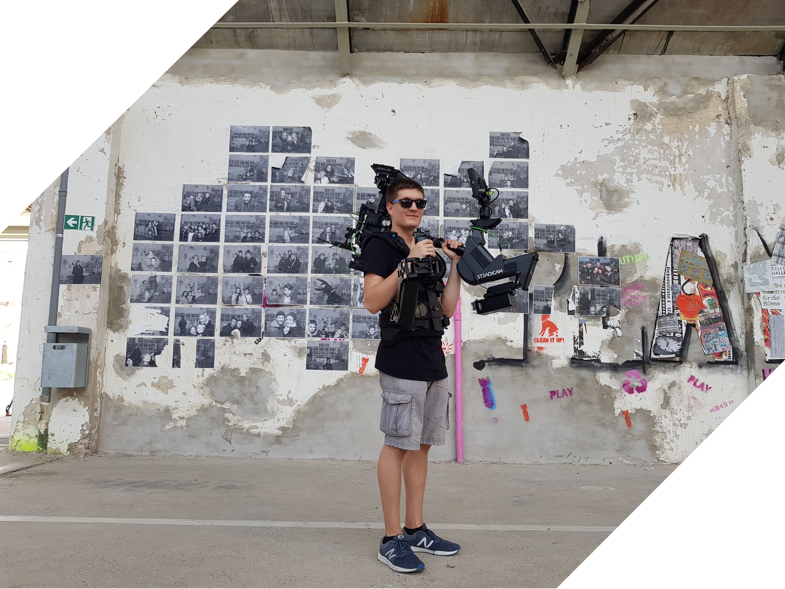 A young man in sunglasses and casual clothing holds professional camera equipment in front of a weathered wall decorated with numerous black-and-white photographs and graffiti.