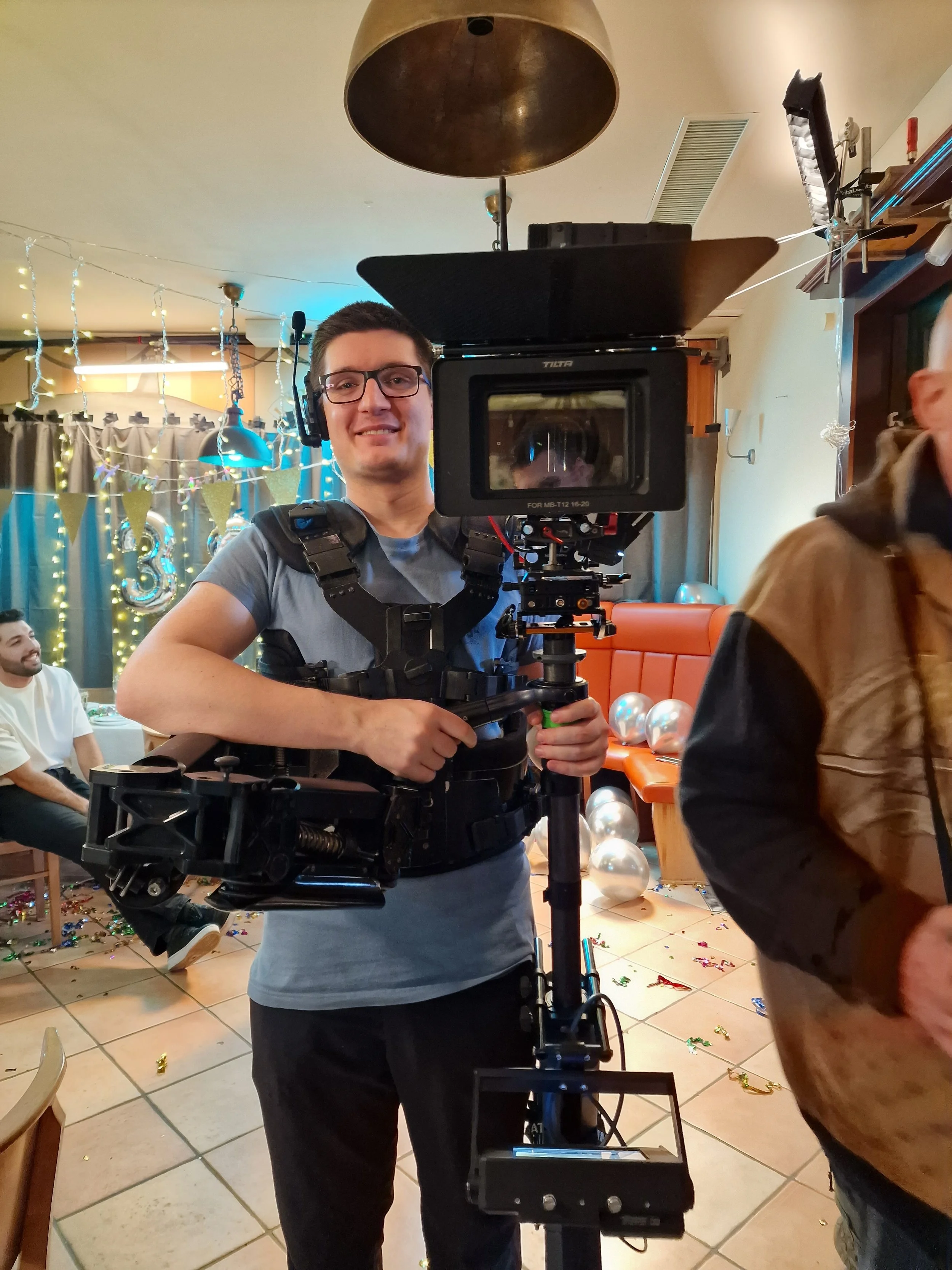 Ein junger Mann mit Brille und grauem T-Shirt, der eine Kamera mit einem Gimbal-Stabilisator in einer festlichen Umgebung bedient, mit bunten Dekorationen und Silvesterballons im Hintergrund.