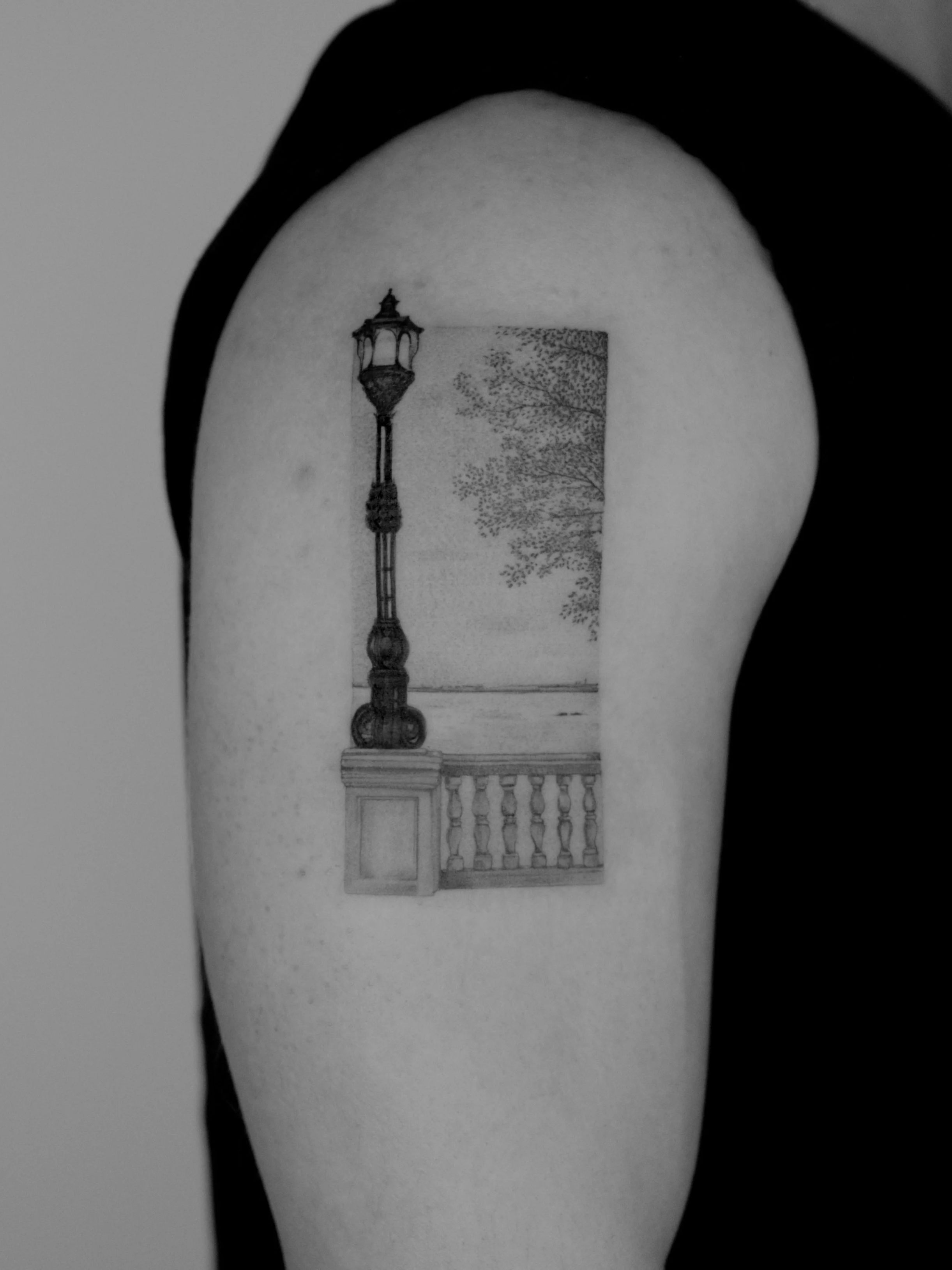 Tatuaje en el brazo de una farola y una baranda de madera con árboles en el fondo.