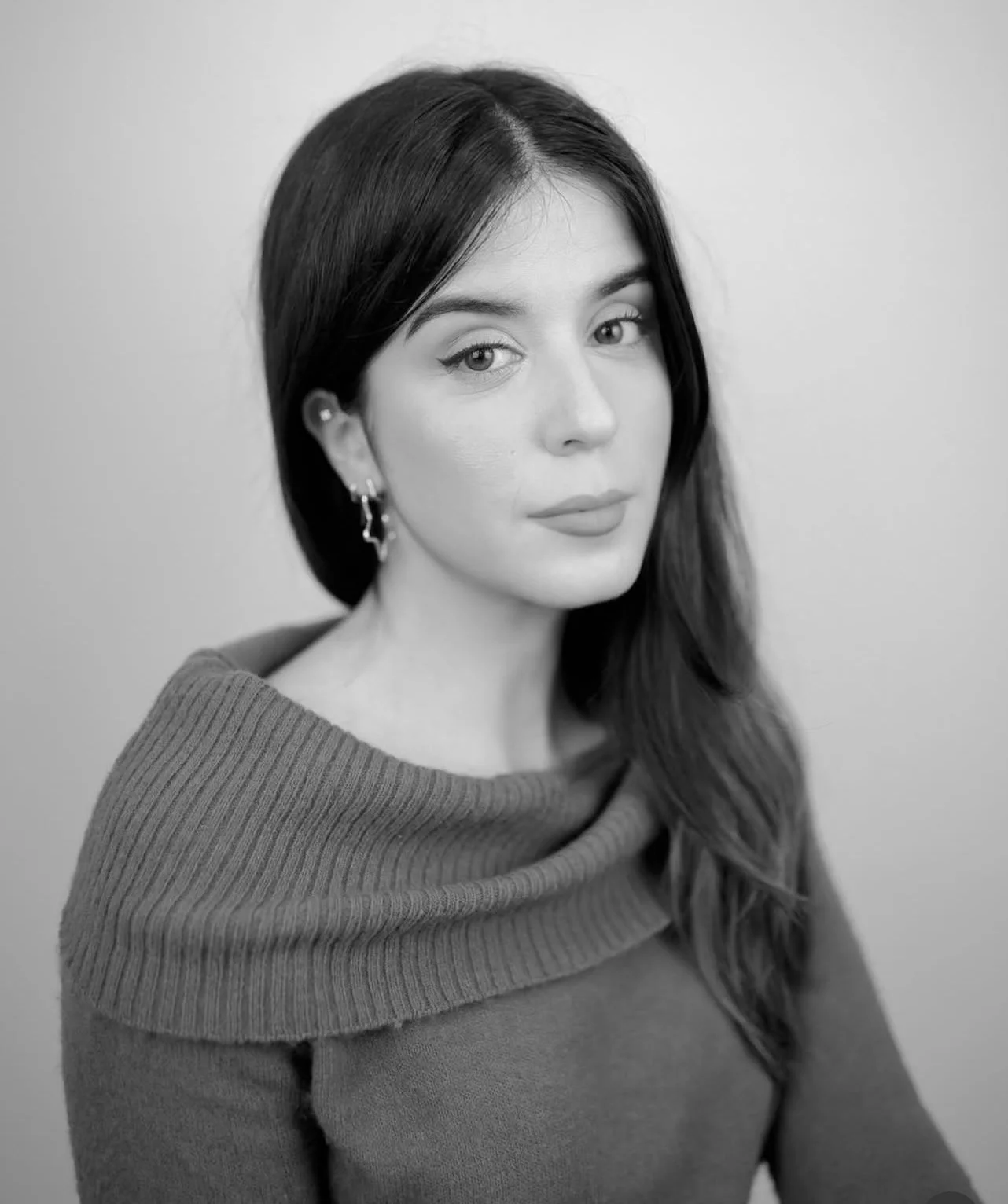 Retrato en blanco y negro de una mujer joven con cabello largo y oscuro, con una expresión serena y ojos claros, usando un suéter de cuello amplio y pendientes pequeños, con fondo sencillo.