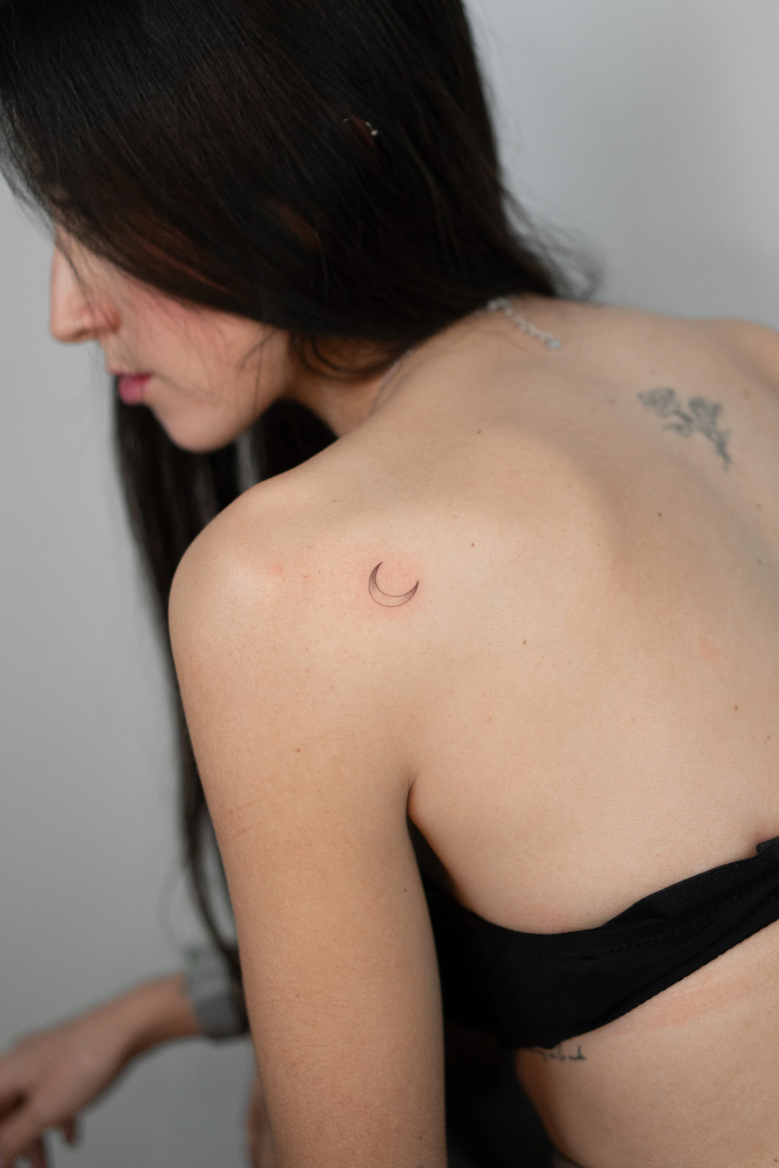 Mujer con cabello largo oscuros, tatuajes en el hombro y el costado, vestida con ropa negra, posando en un fondo gris.