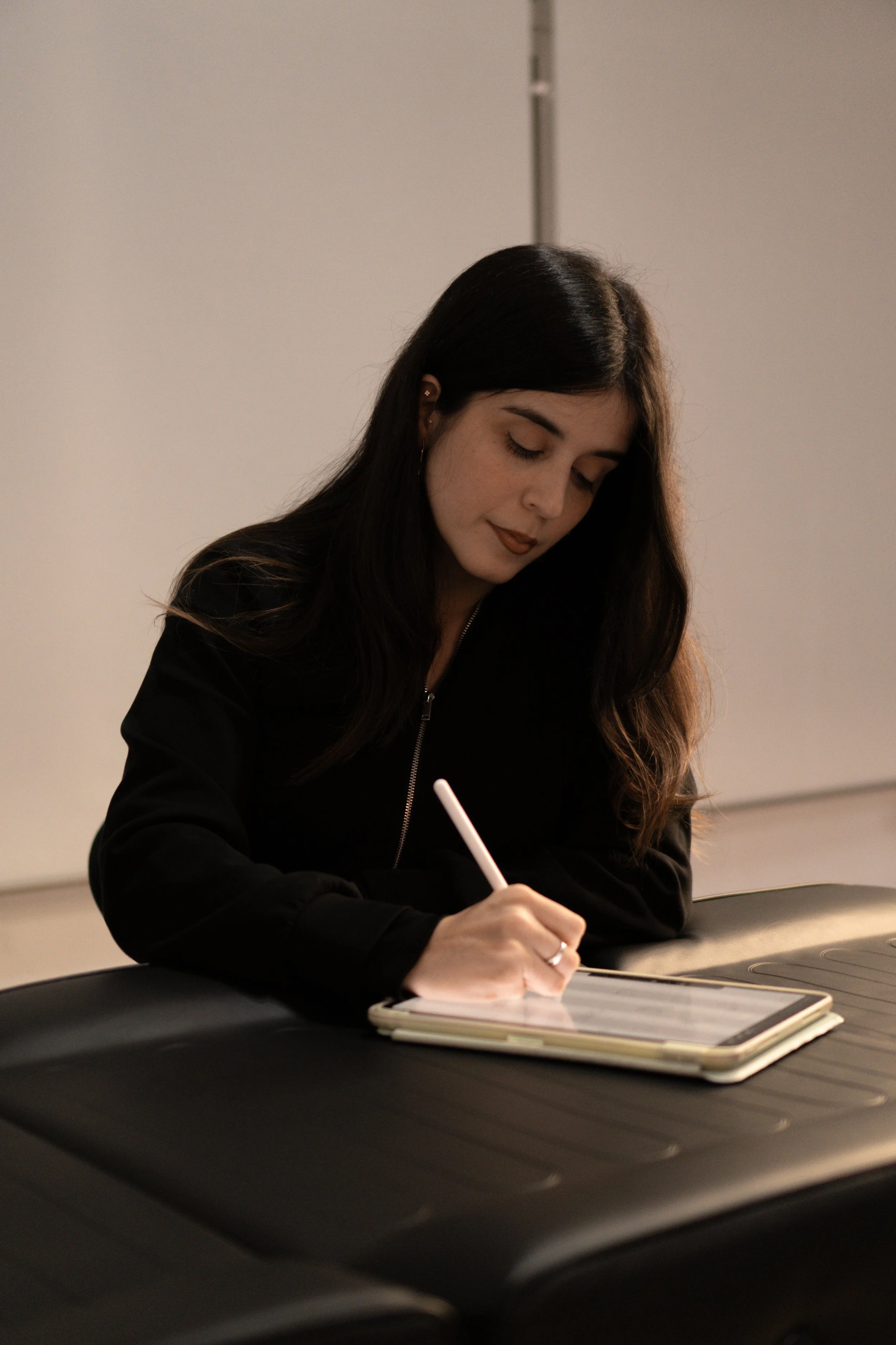 Mujer con cabello largo y oscuro, maquillada, escribiendo en un bloc de notas en una mesa, usando ropa negra, en un ambiente interior de oficina o sala de reuniones.