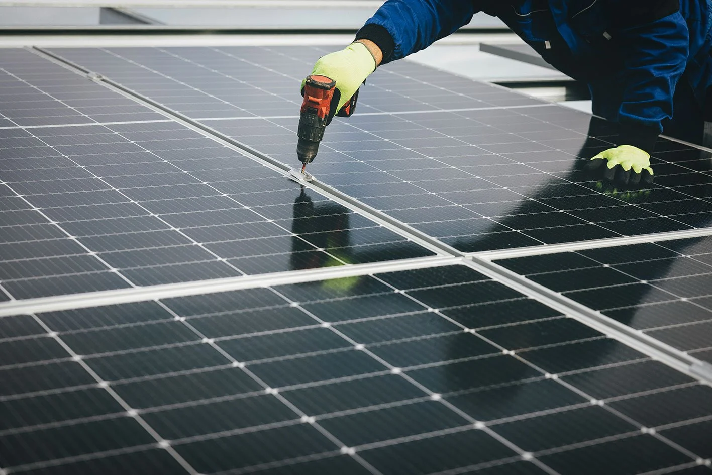 Worker installing or maintaining solar panels using a cordless drill, wearing gloves and a blue jacket.