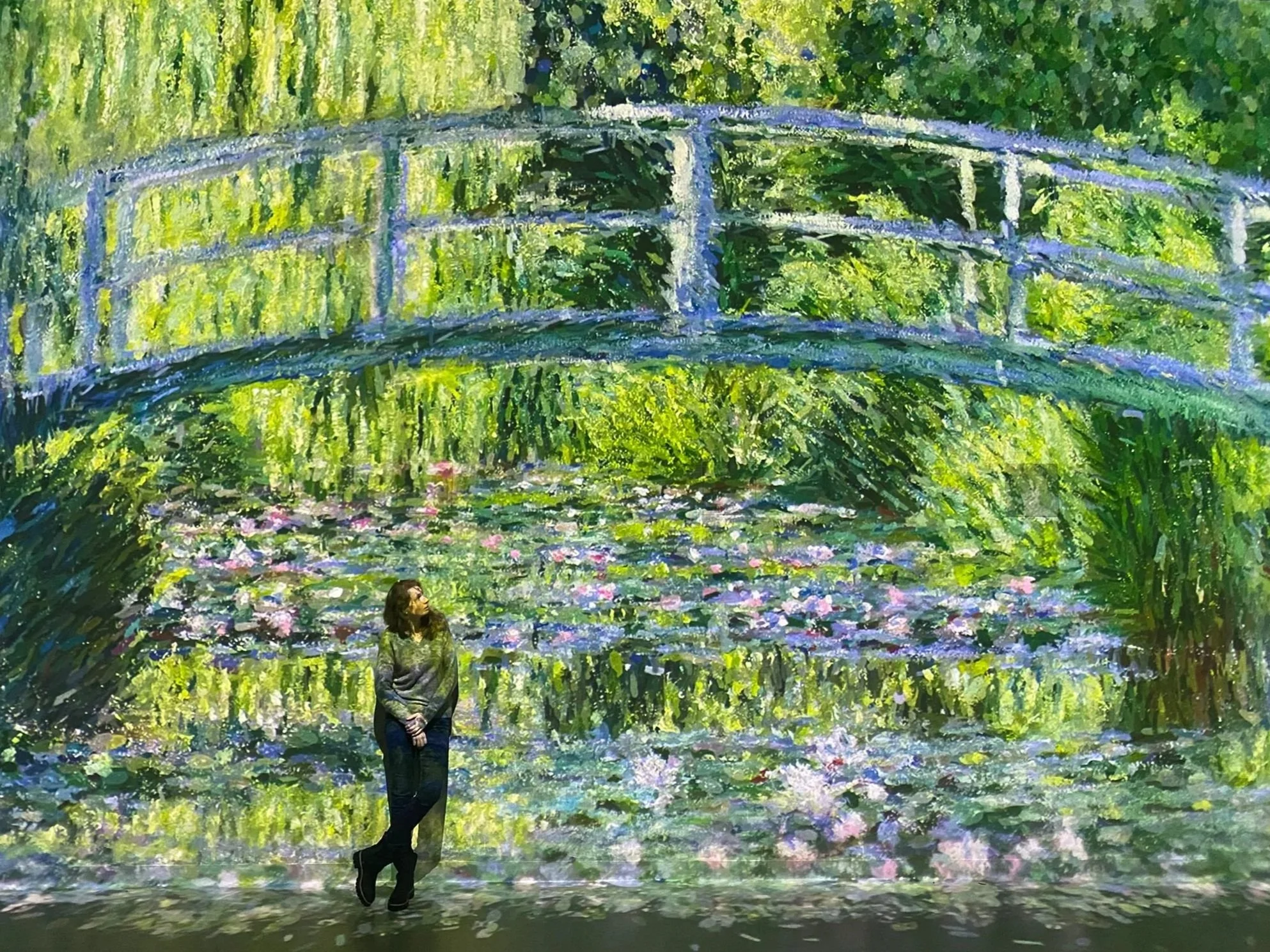 A woman is standing in front of a large animated mural of a water scene, including a bridge over water with pink and purple flowers and lush green trees and foliage.