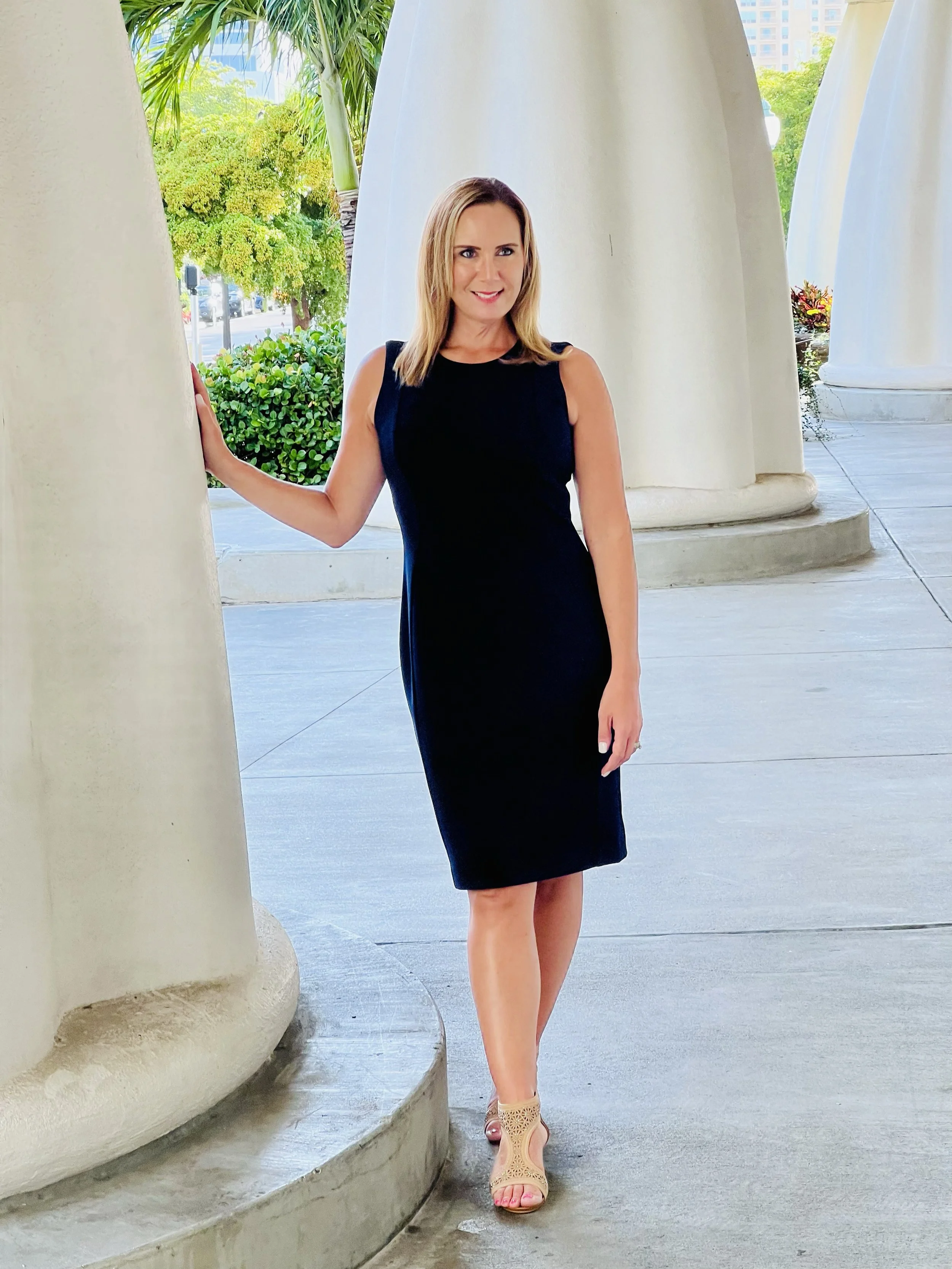 A woman standing outdoors near columns, wearing a black sleeveless dress and beige open-toe heels, with a smile, greenery, and urban buildings in the background.