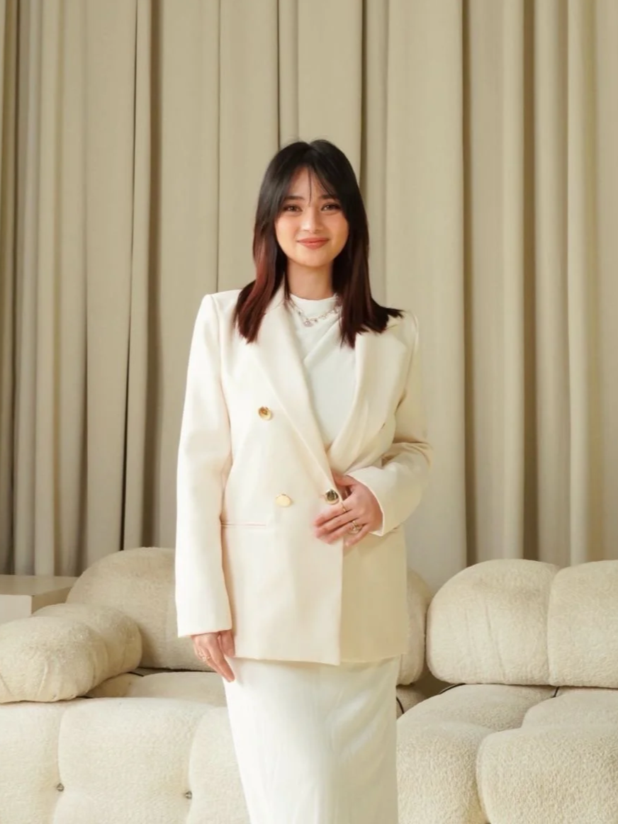 A woman standing in a cream-colored room with beige drapes and a cream sofa, wearing a white blazer, white dress, and jewelry, smiling at the camera.