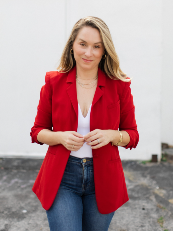 Woman wearing a red blazer, white top, and jeans, standing outdoors against a plain white wall.