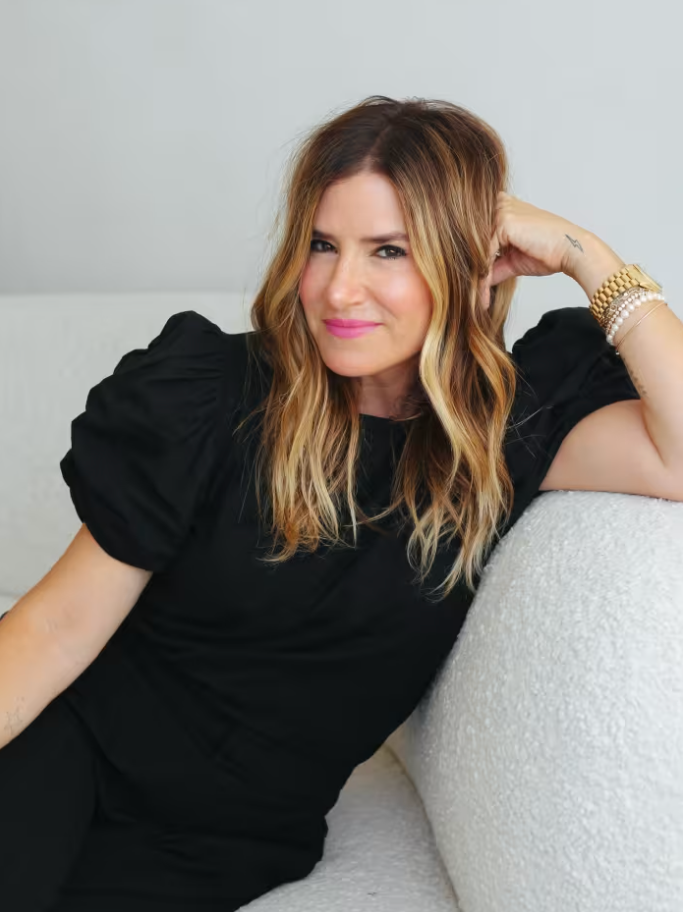 A woman with wavy blonde hair and pink lipstick sitting on a white couch, wearing a black top and bracelets, looking at the camera.