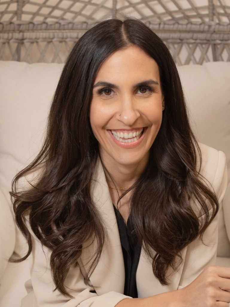 A woman with long dark hair and fair skin, smiling at the camera, wearing a beige blazer over a black top, sitting against a neutral background.