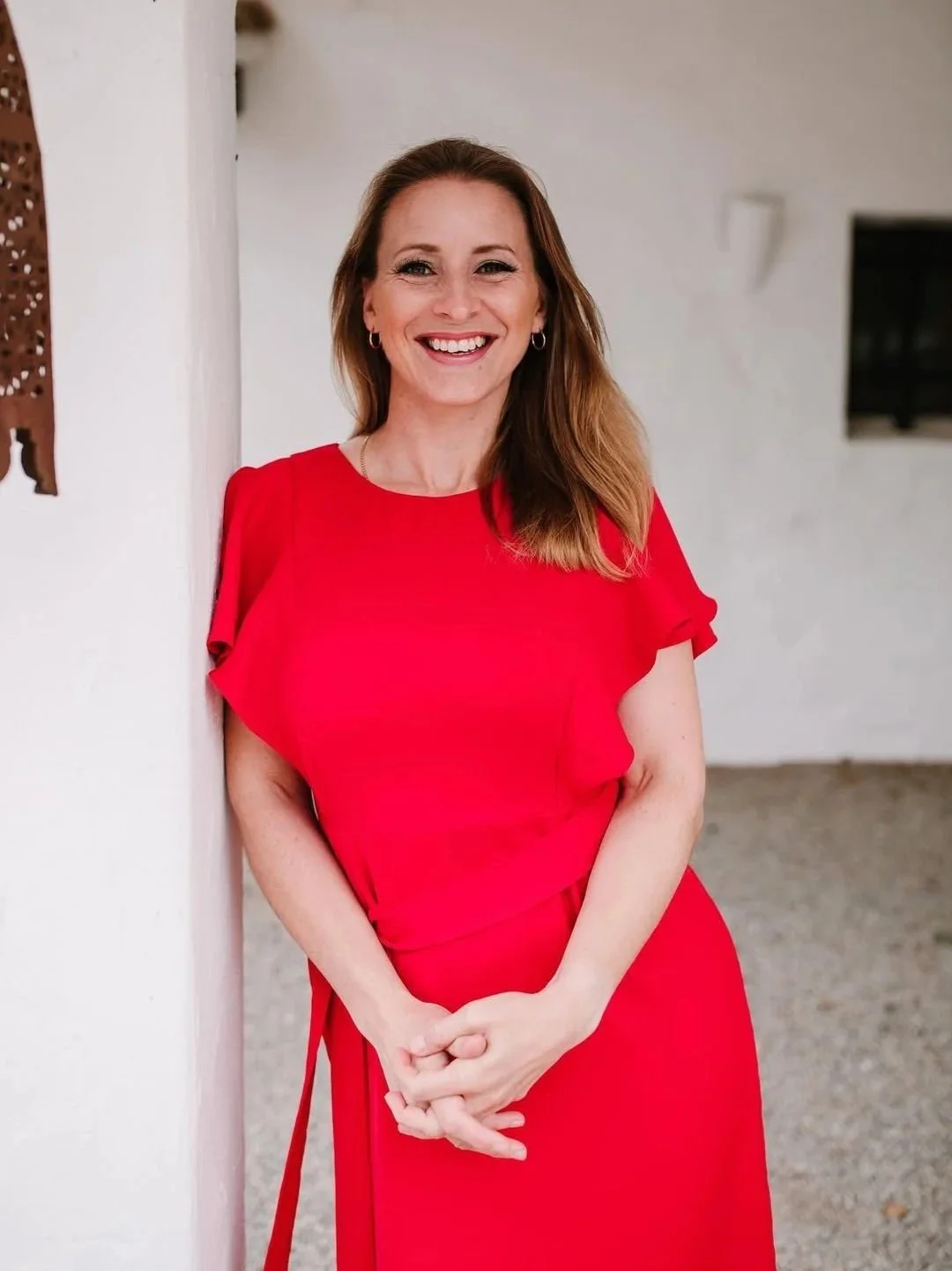 Woman in red dress smiling and standing against a white wall.