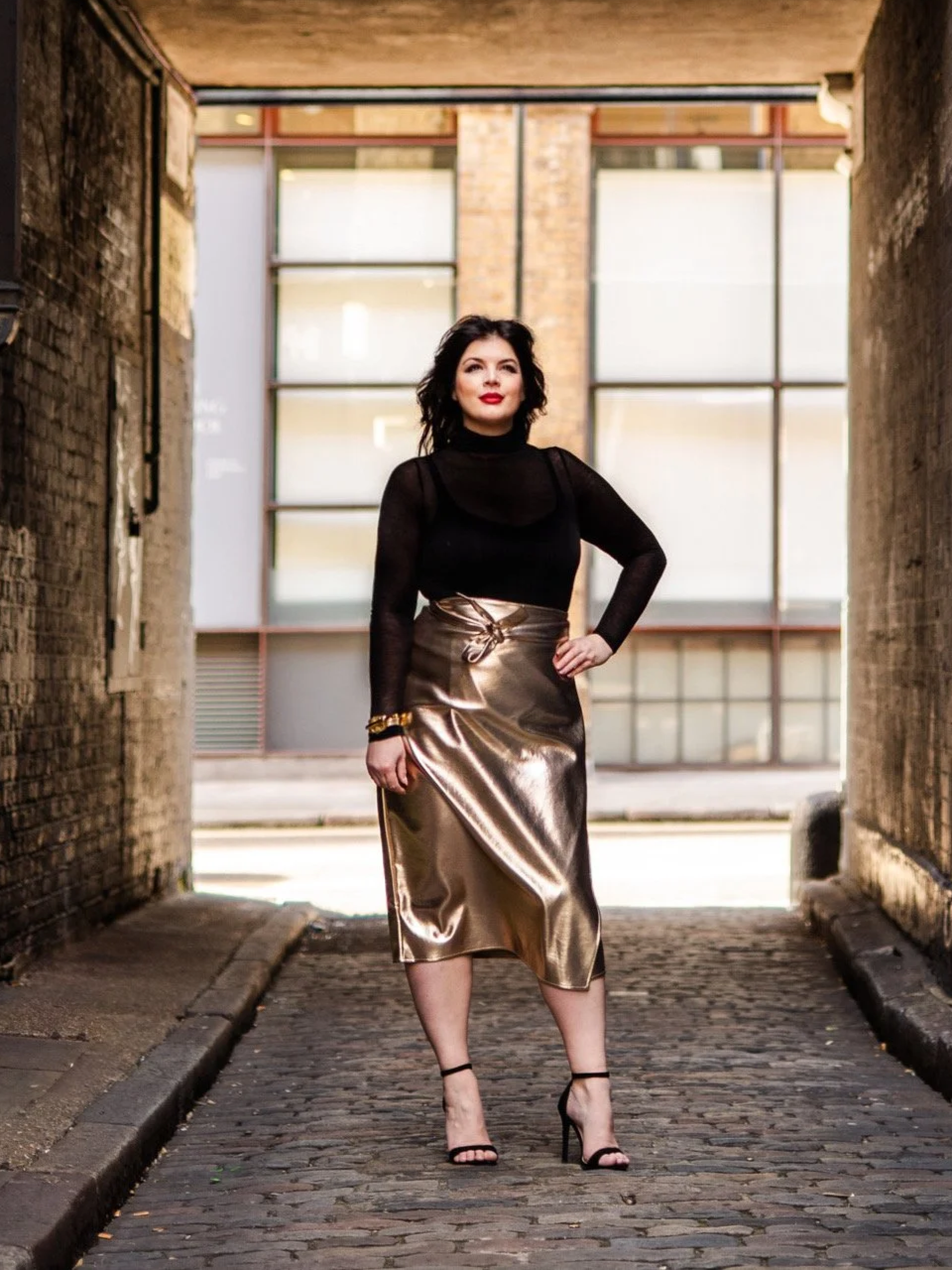 A woman standing in a narrow alleyway with brick walls, wearing a black long-sleeve top, a shiny gold midi skirt, and black high heels.