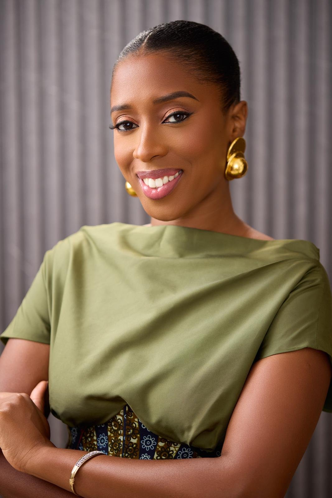 A woman with dark skin smiling, wearing large gold earrings, a green satin top, and a patterned skirt, standing in front of a gray curtain.